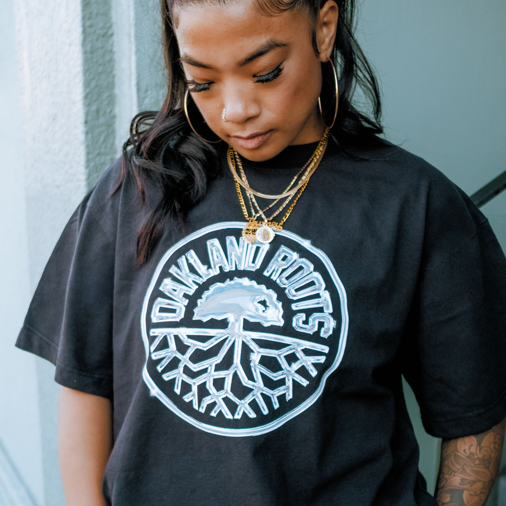 A woman with hoop earrings and layered gold necklaces looks down, partially revealing her tattooed arm. She wears the Oakland Roots SC Platinum Crest Tee, featuring a large white tree logo with roots inside a circle.