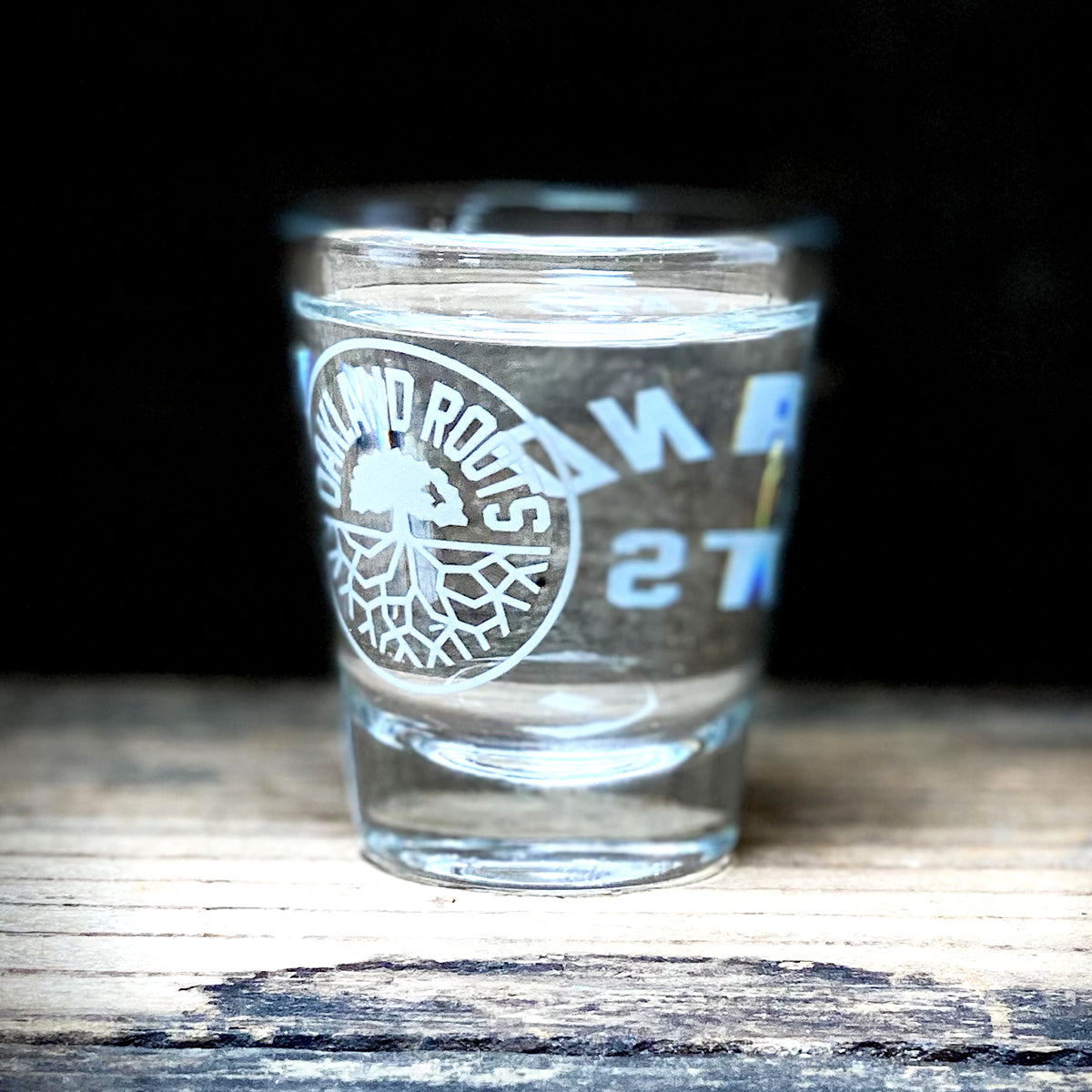 An Oakland Roots SC Shot Glass rests on a wooden surface against a dark background. The front of the glass displays a white logo with "DIAMOND ROOTS" encircling an image of a tree with spreading roots. The empty glass, though slightly out of focus, reflects the strong community spirit of Oakland Roots Sports Club.