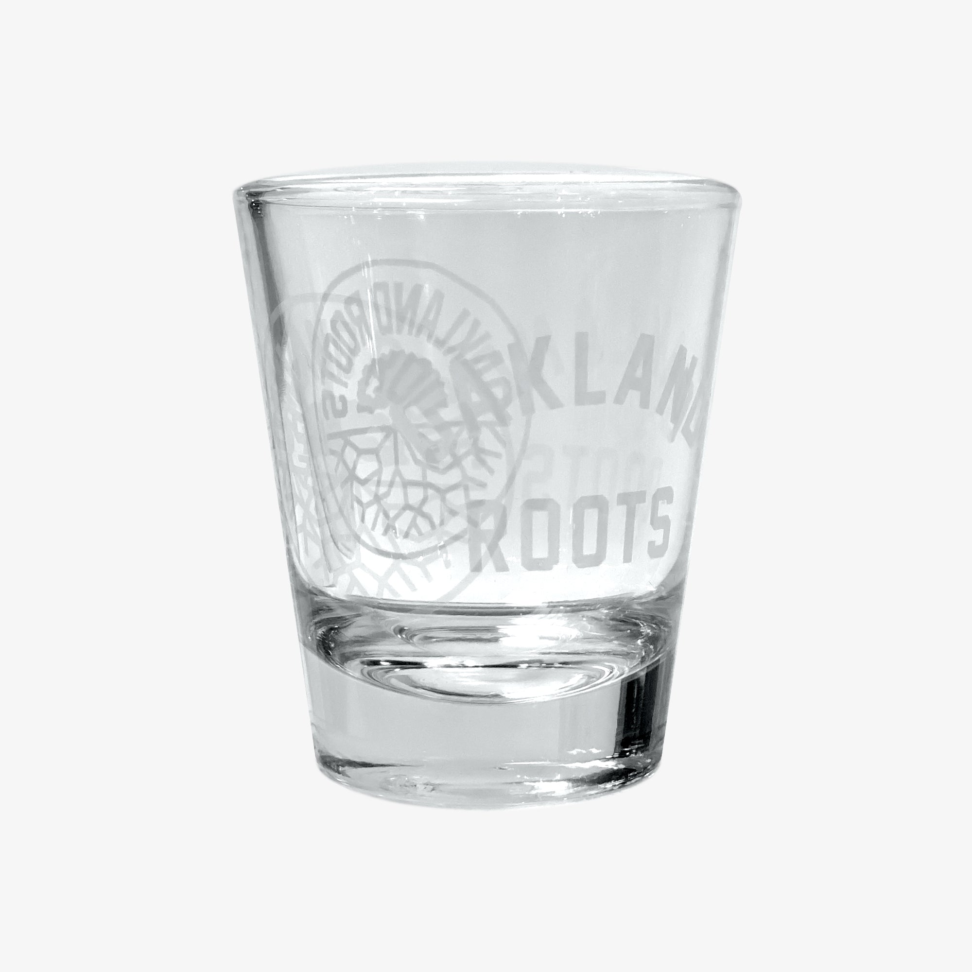 The Oakland Roots SC Shot Glass from Oakland Roots SC is a clear glass with a wide rim that tapers towards the base. It boasts an etched design featuring a detailed circular emblem and the words "OAKLAND ROOTS SPORTS CLUB" prominently displayed. With its plain white background, this shot glass is perfect for any Oakland fan.