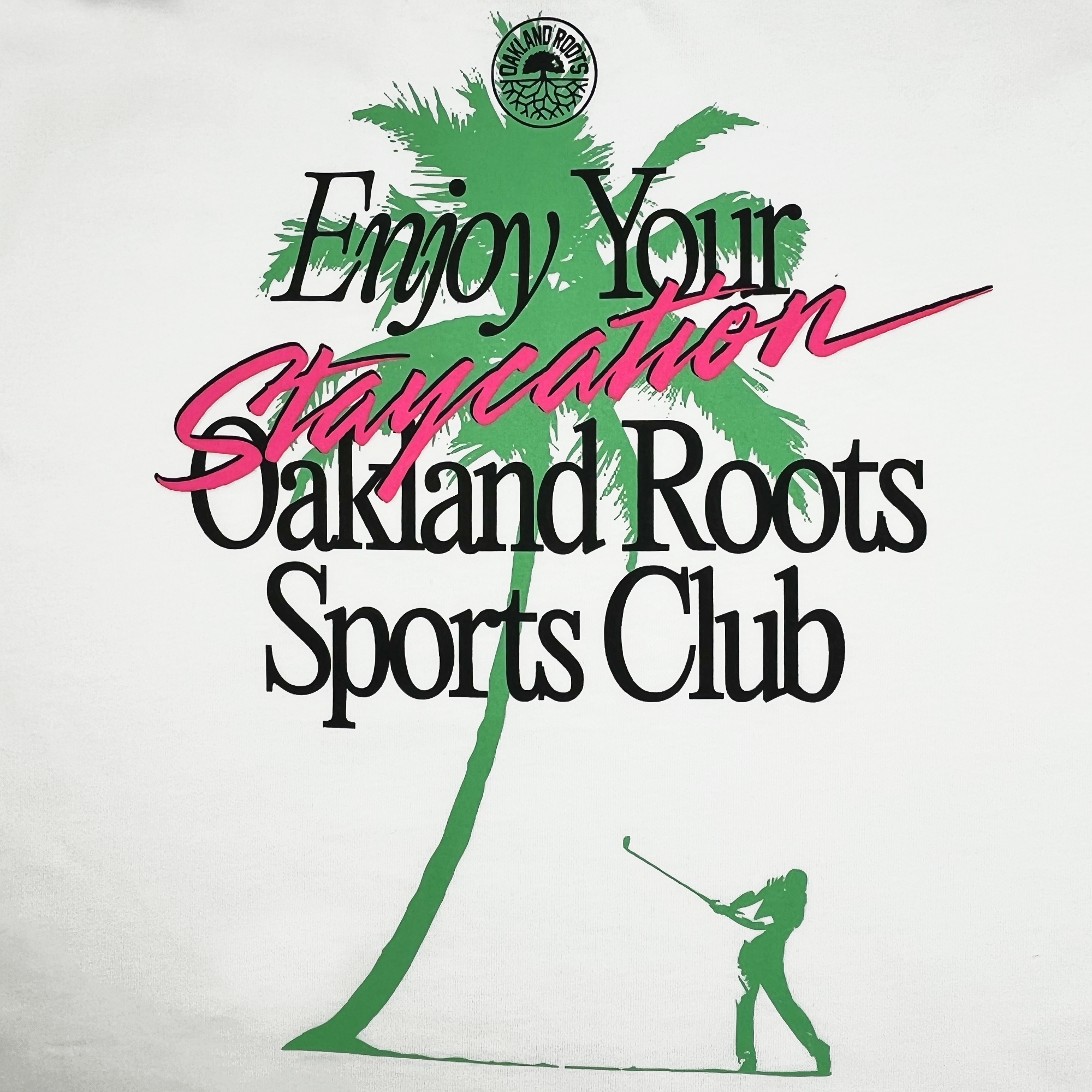 A graphic showcases a green silhouette of a palm tree and a person swinging a golf club, with overlay text that reads "Enjoy Your Staycation" in cursive, and "Staycation" highlighted in pink. Below it says "Oakland Roots SC" in black. The small circular logo appears in the upper left corner, making it perfect for the Oaklandish Boxy fit Staycation Heavy Tee from Oakland Roots Sports Club promoting social good.