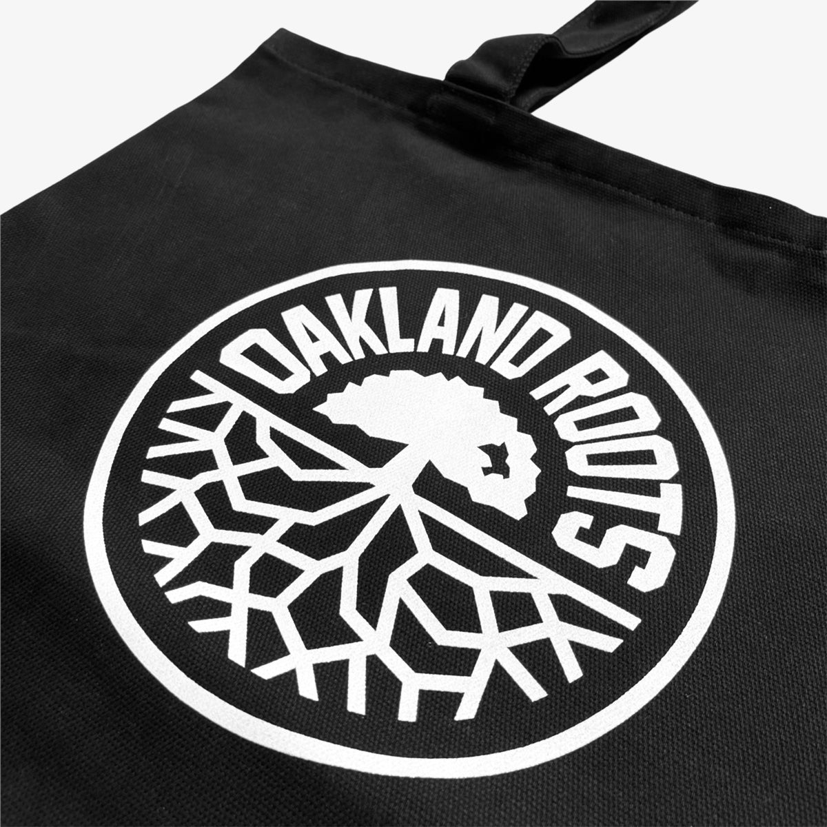 The Oakland Roots SC Tote from Oakland Roots SC showcases a striking white circular logo against its black fabric. The design features bold text spelling 'Oakland Roots' encircling a tree with branching roots, capturing the essence of an Oaklandish vibe. It also includes a single black strap for easy carrying.