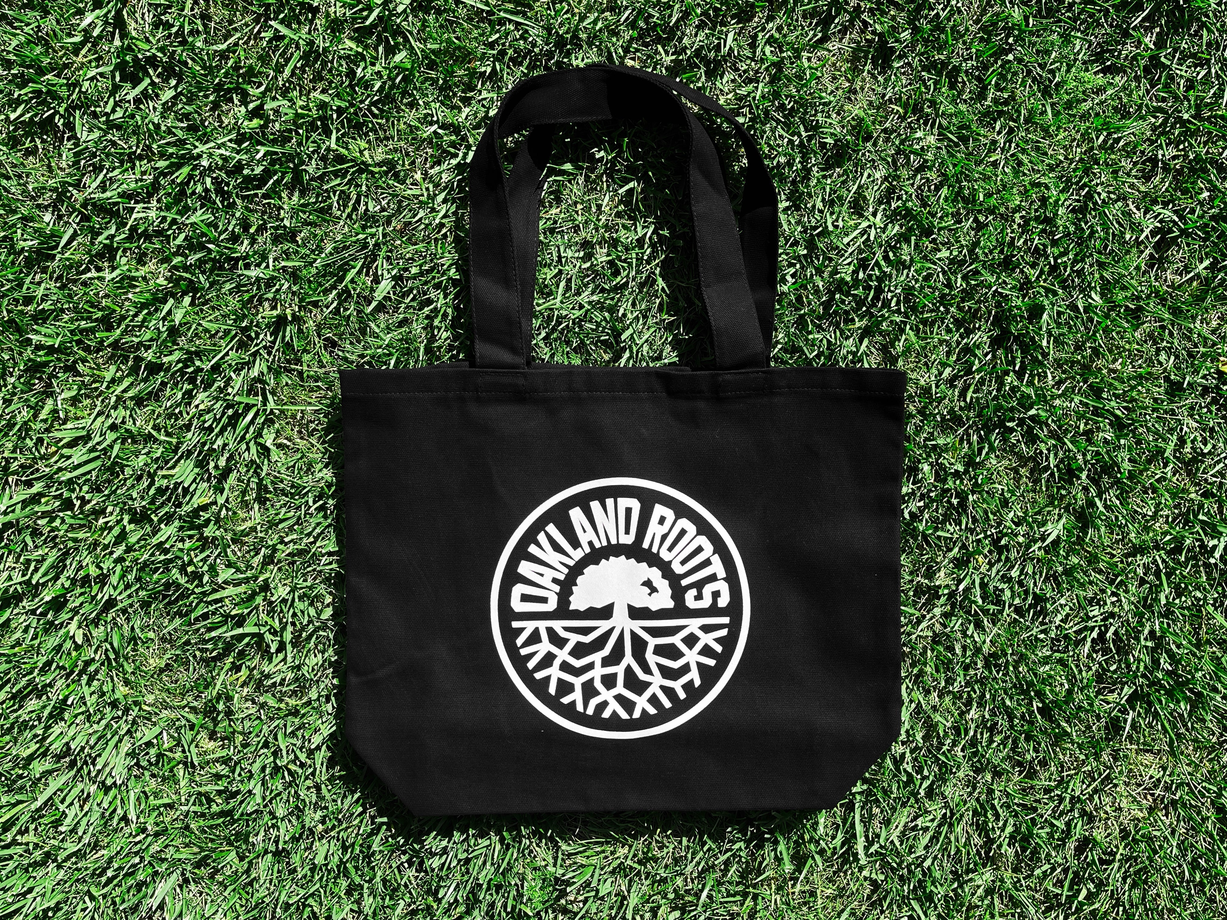 An Oakland Roots SC Tote is placed on grass. The black bag, featuring two handles, showcases a white circular logo displaying an oak tree with roots graphic. The text "OAKLAND ROOTS" encircles the tree within the logo, symbolizing its ties to the Oaklandish heritage and local sports pride. The tote casts a slight shadow on the grass.