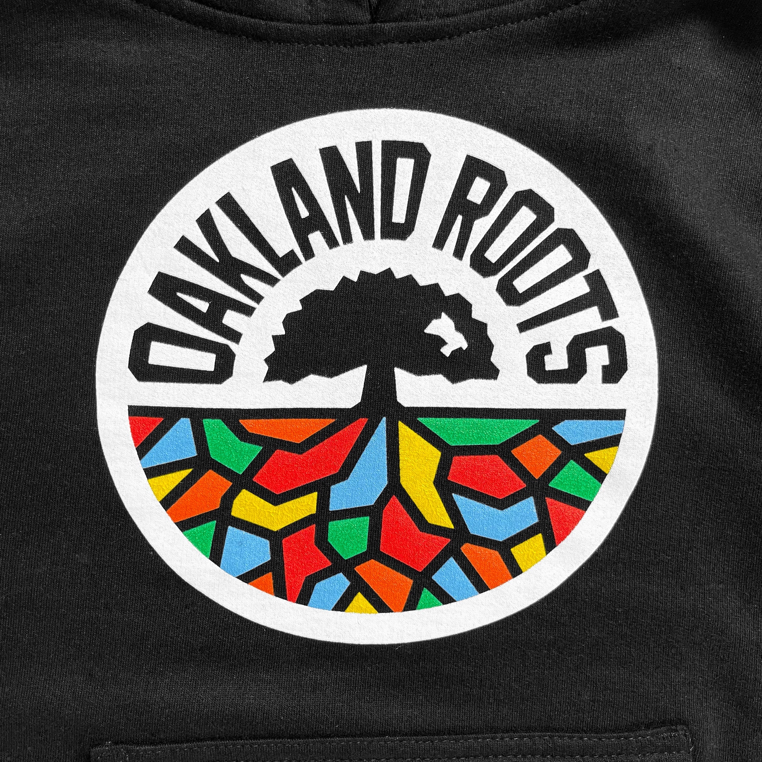 A Youth Oakland Roots SC Hoodie showcases a circular "Oakland Roots" logo on a black background. The logo features a black tree silhouette reminiscent of the Oaklandish design, positioned above colorful geometric shapes in red, yellow, green, and blue that resemble roots. These elements are all enclosed within a white circle with the text "OAKLAND ROOTS" arching over its top edge.