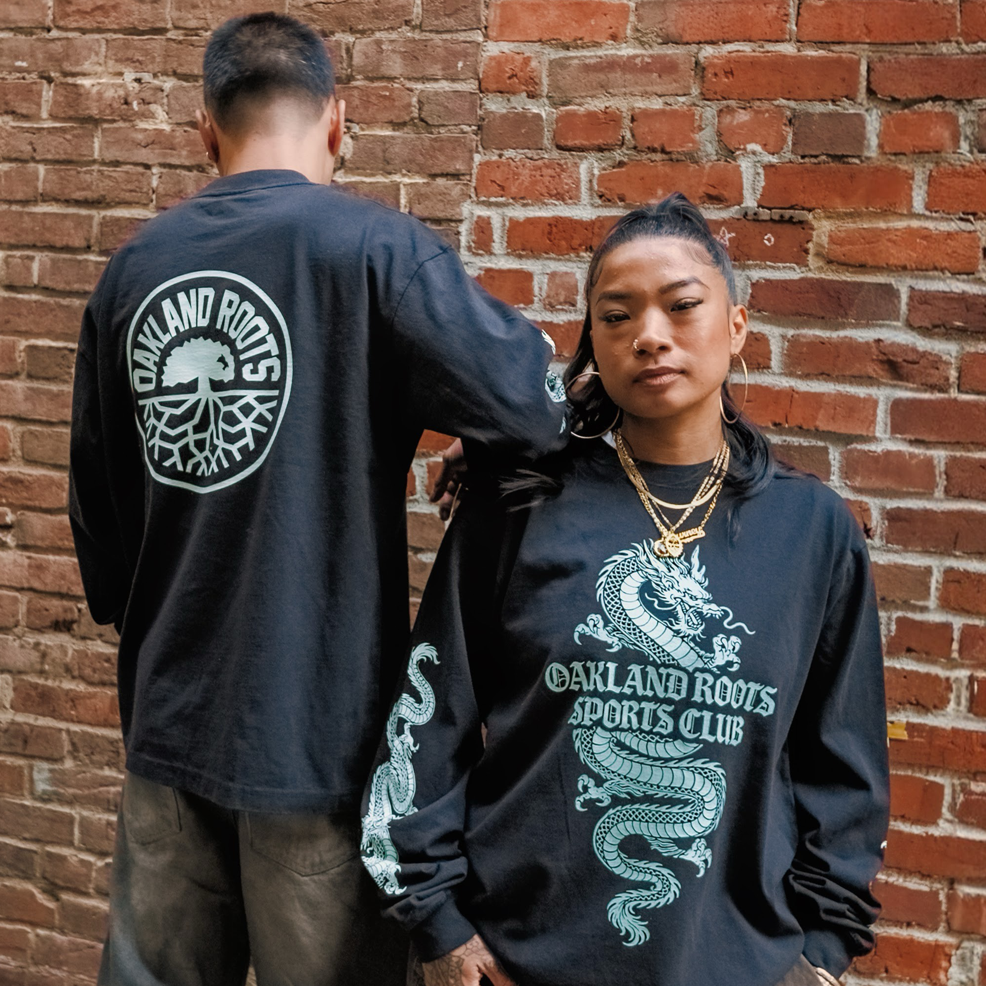 Two people pose confidently against a brick wall in matching black Oakland Roots SC ORSC Ryujin LS Tees, each featuring a white dragon design and the iconic roots logo on the back—showcasing bold style from Oakland Roots SC.