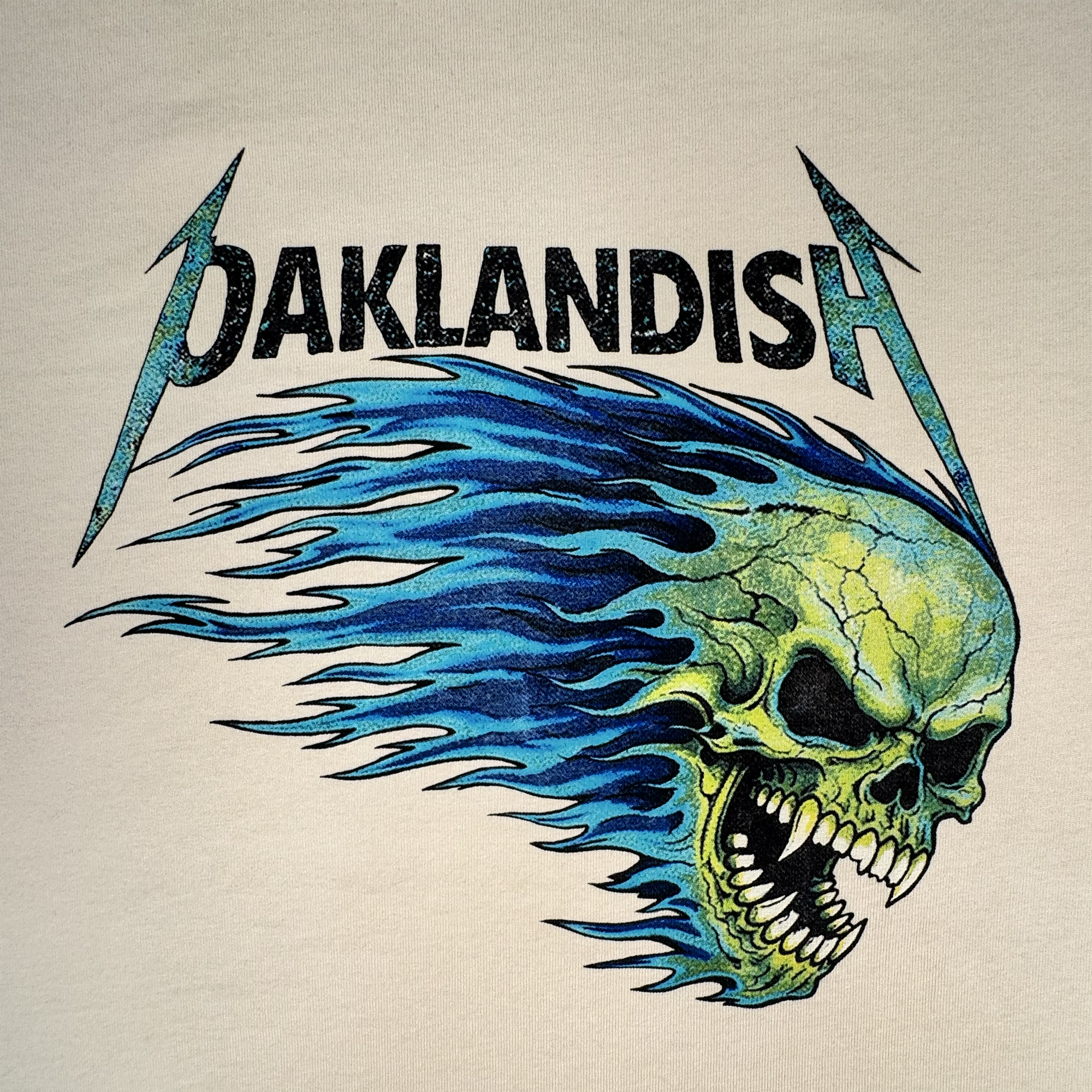 Sure! Here’s a rewritten version using your product data: The Sandman Tee by Oaklandish features bold black “Oaklandish” lettering with electric blue lightning bolts. Below the text, a vivid green skull with streaming blue flames is shown facing left, mouth open and baring sharp teeth with hollow eyes. Printed on a classic fit t-shirt, this striking design pops against the plain beige background.
