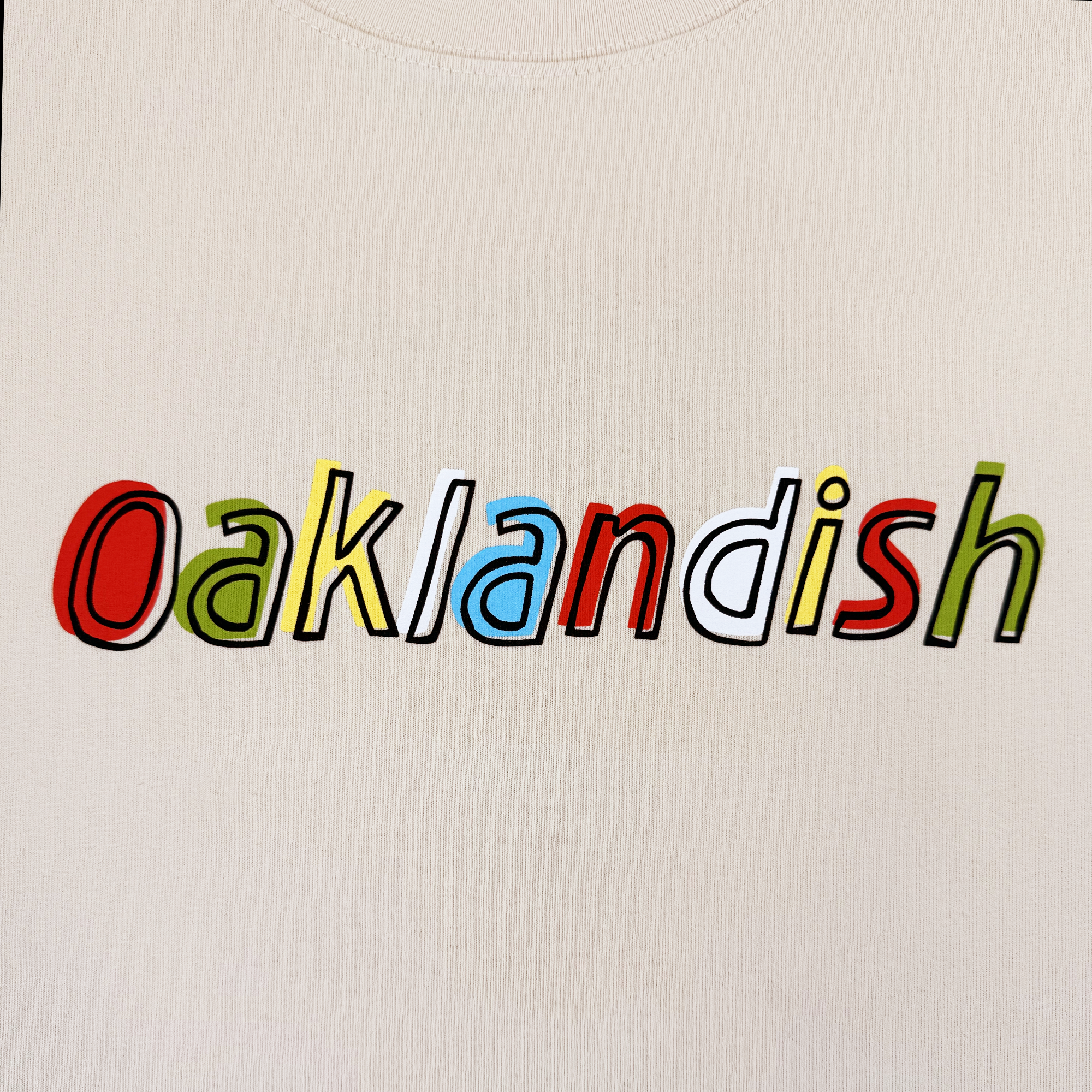 The Oaklandish Scrawl Tee features “Oaklandish” in colorful, overlapping letters outlined in black on a cream heavyweight cotton men’s shirt, blending red, green, yellow, blue, and white for a bold and playful look.