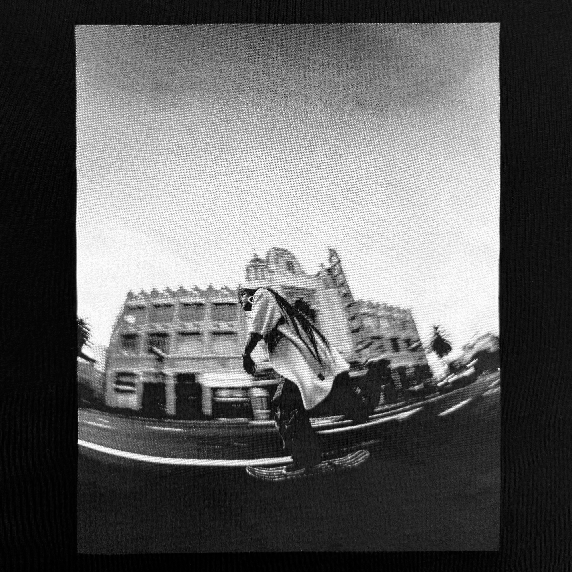 The "Skateboarding is Beautiful Tee" by Oaklandish features a striking black and white image of a skateboarder captured mid-motion through a fisheye lens, highlighting the artistry of skateboarding. The photograph focuses on the skater, set against the backdrop of an ornate historic building and blurred palm trees along an Oakland street.