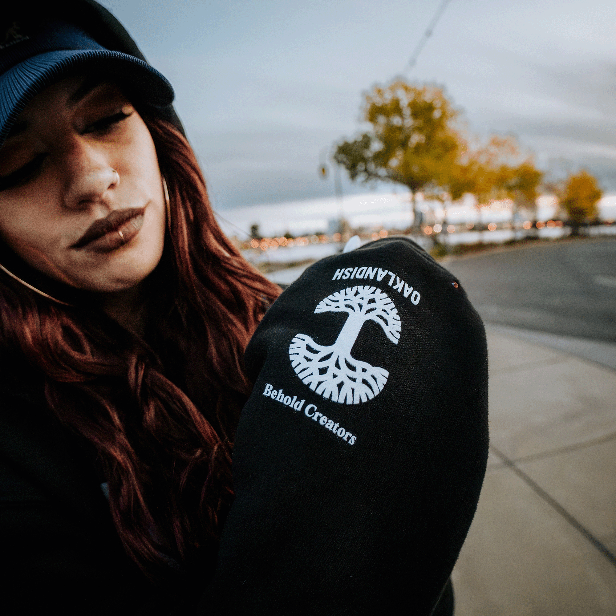 A person with long red hair, sporting a black cap and the "Skateboarding is Beautiful Hoodie" from Oaklandish, which features a white tree design along with the words "OAKLANDISH" and "Behold Creators," perfectly captures the lively essence of Oakland. Surrounded by city streets, yellow-leaved trees, and a cloudy sky, this scene invites the eye of an urban photographer.