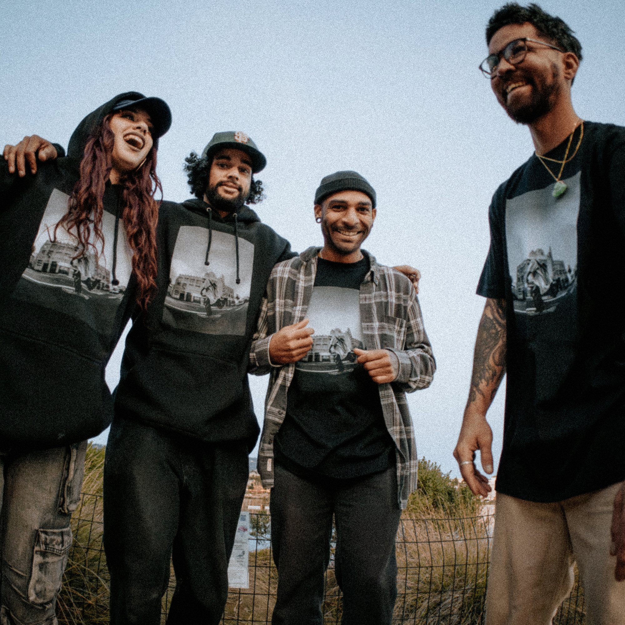 A group of four people outdoors in Oakland, smiling and standing together. Three are wearing matching black shirts featuring the same design, while one is sporting an "Oaklandish" brand hoodie called "Skateboarding is Beautiful." The clear sky and blurred greenery hint at a casual gathering. Their relaxed demeanor suggests they might be skaters or friends capturing the moment with a photographer.