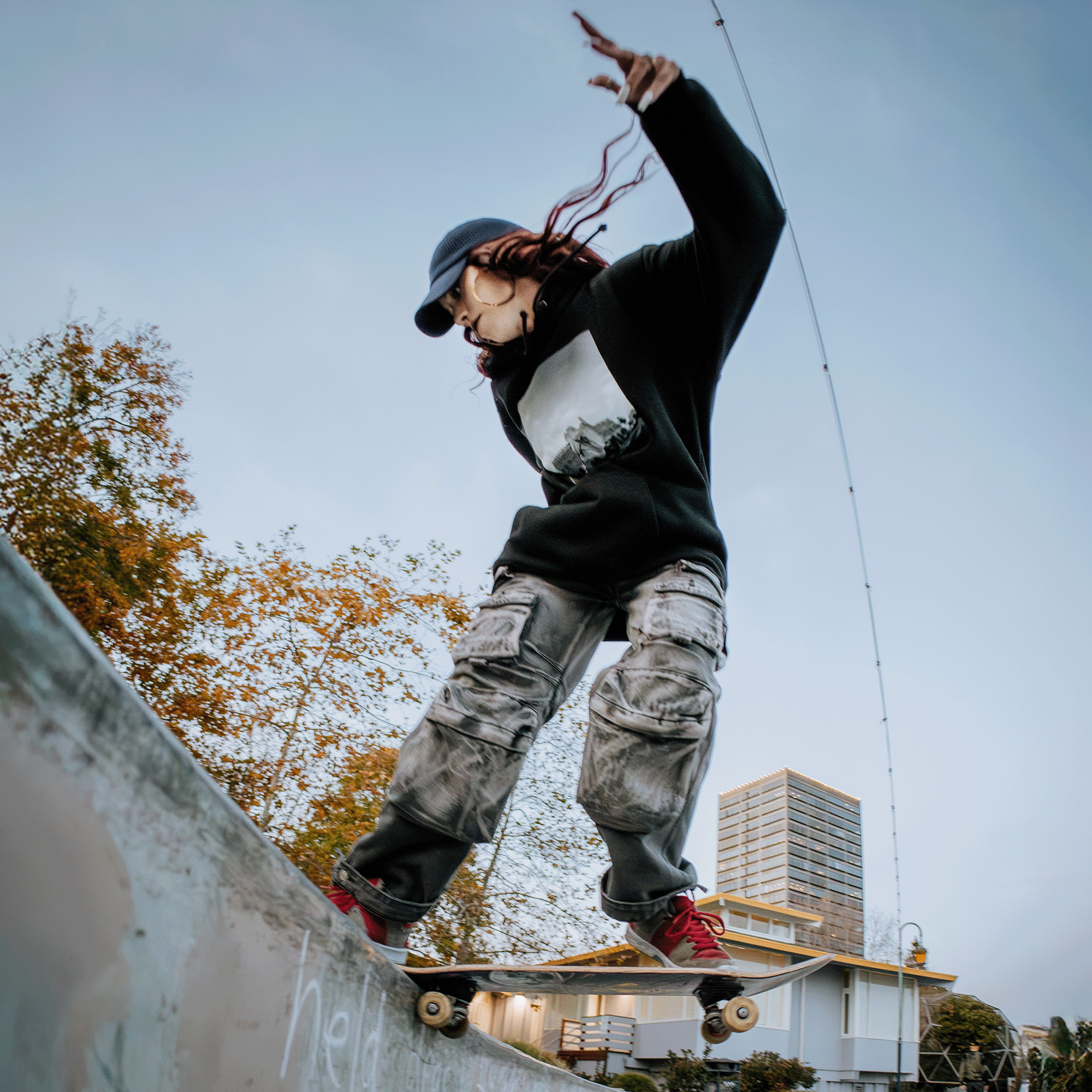 A skateboarder with long hair and a cap performs a trick on a concrete ledge in Oakland. They wear an Oaklandish "Skateboarding is Beautiful" hoodie, gray cargo pants, and red sneakers. A photographer captures the moment as trees and a tall building stand in the background under the clear blue sky.