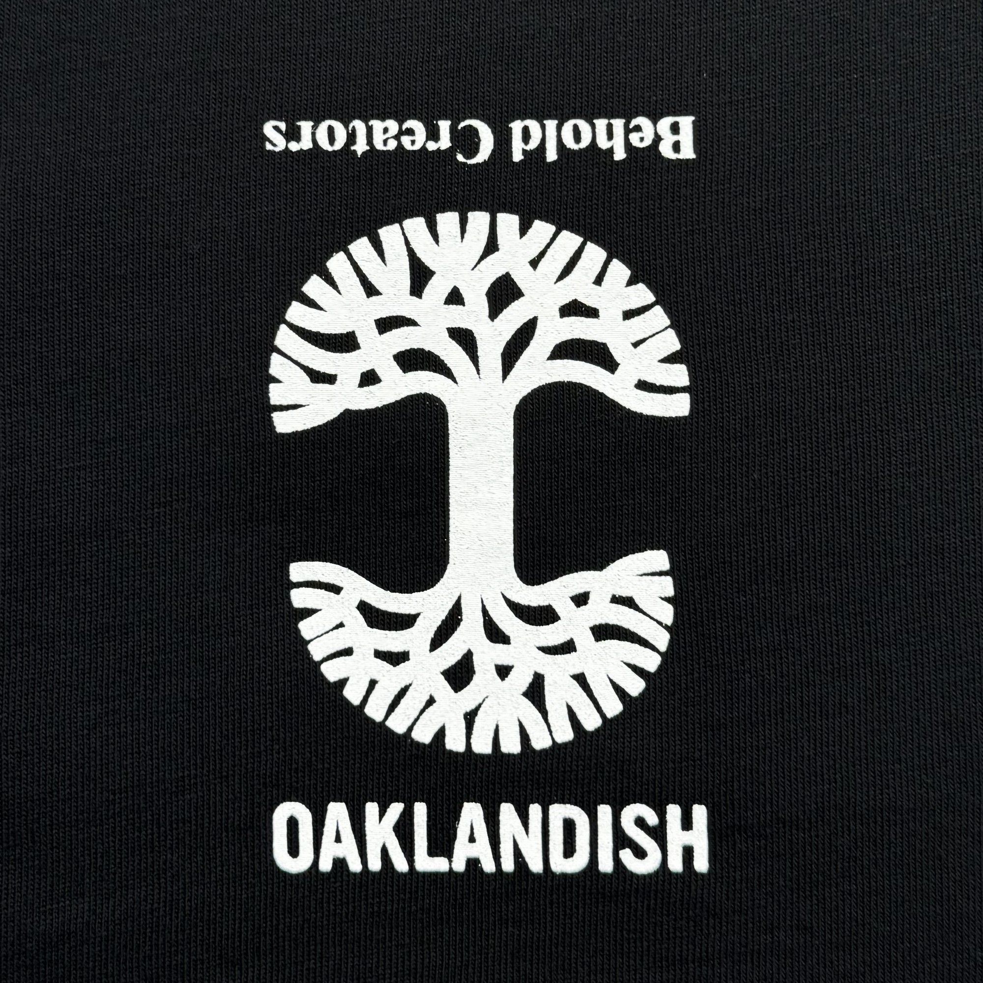 The Skateboarding is Beautiful Tee from Oaklandish features a striking black background with a white illustration of a tree and symmetrically extending roots in a circular design. Beneath the tree, "OAKLANDISH" is boldly printed in white capital letters, while "Betond Creations" appears upside down above it, encapsulating the vibrant spirit of Oakland and its skateboarding culture.