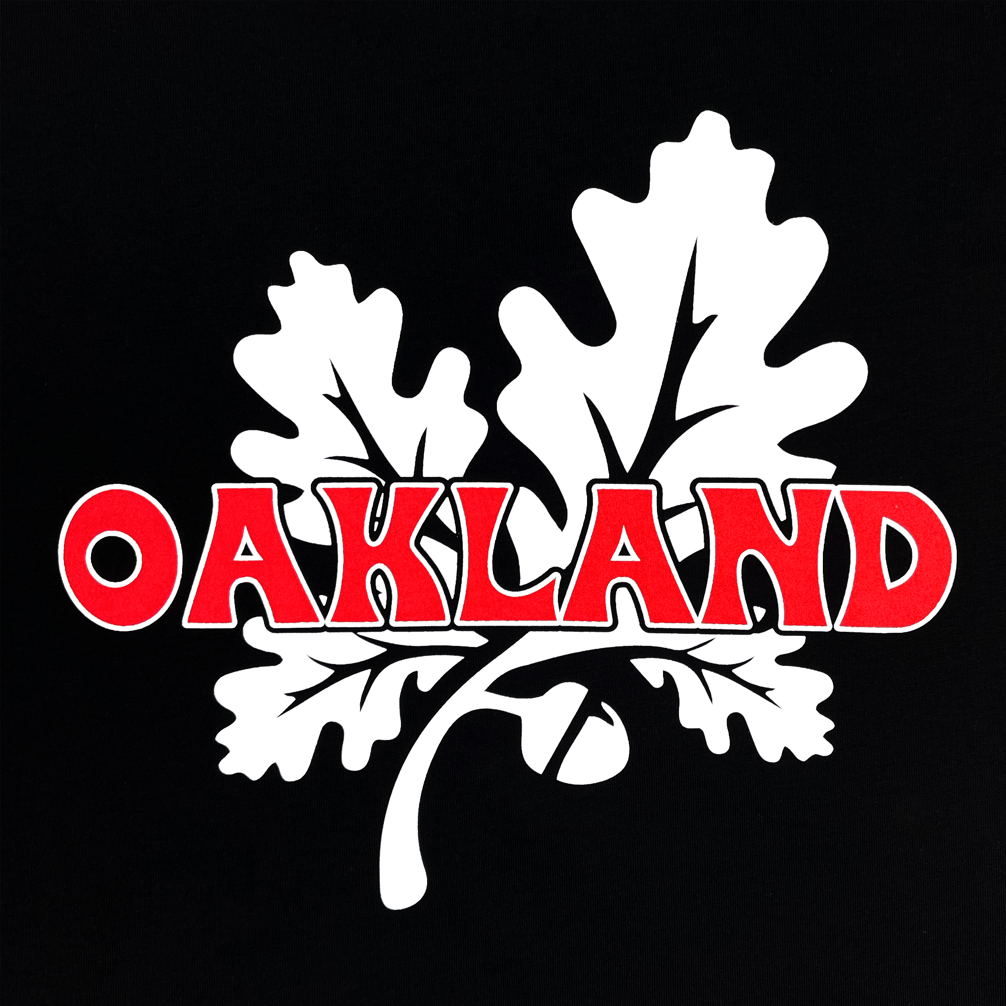 An illustration of bold, red "OAKLAND" text with white borders is placed above and through white oak leaves on a black 100% cotton classic fit t-shirt. The oak leaves are detailed with black veins, and there is an acorn attached to the stem on the left side, featured on Oaklandish's Grocery Run Tee.