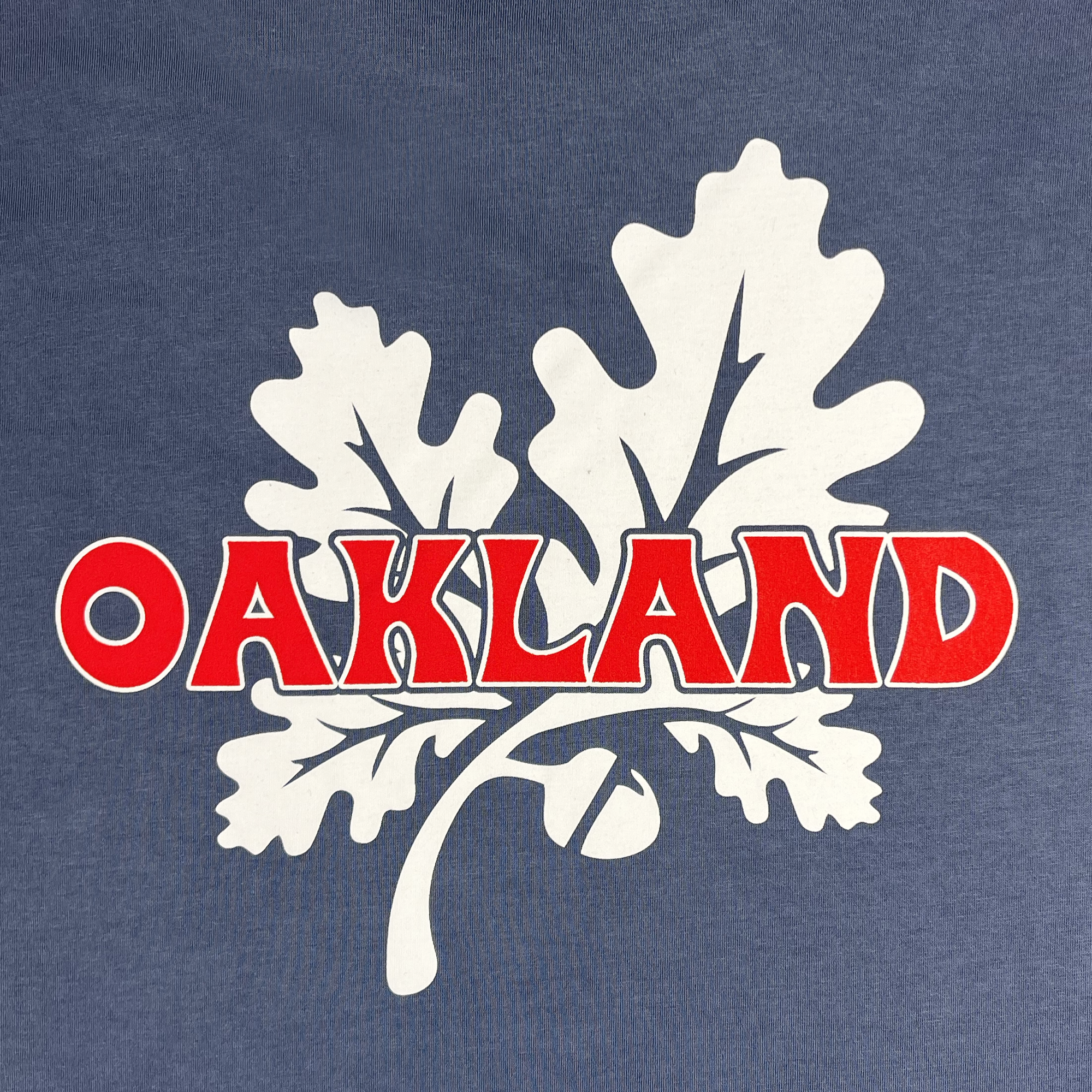 Introducing the Grocery Run Tee by Oaklandish: This classic fit t-shirt showcases a graphic design featuring the word "OAKLAND" in bold, red uppercase letters with a white outline. Two white oak leaves and an acorn sit below the text, all set against a grayish-blue background. Made from 100% cotton, this tee ensures both comfort and style.