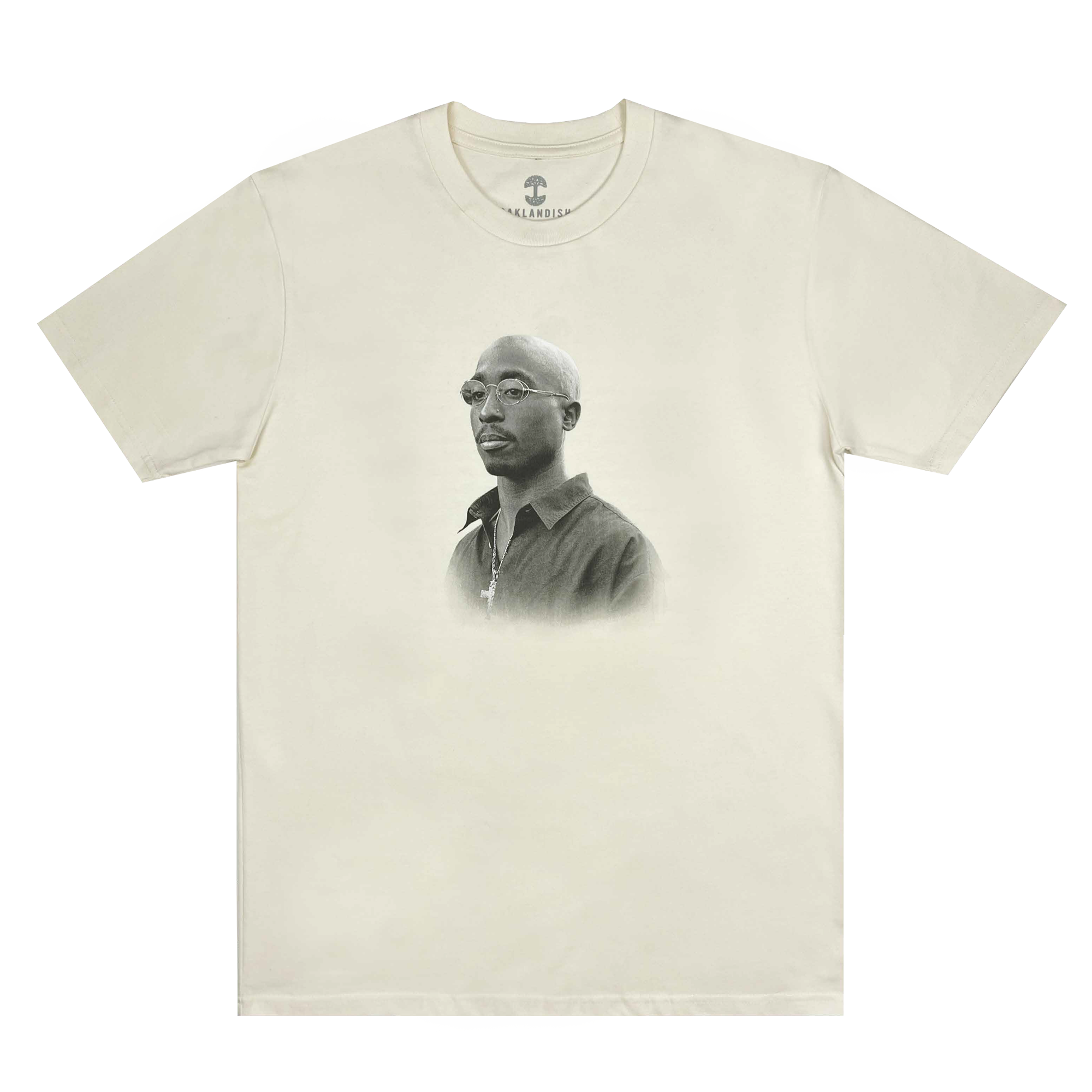 The Sometimes Eye Cry Tee by Oaklandish showcases a light beige design with a black and white portrait of Tupac Shakur in glasses and a button-up shirt prominently displayed on the chest. This short-sleeved, round-neckline T-shirt captures the timeless Oaklandish style.