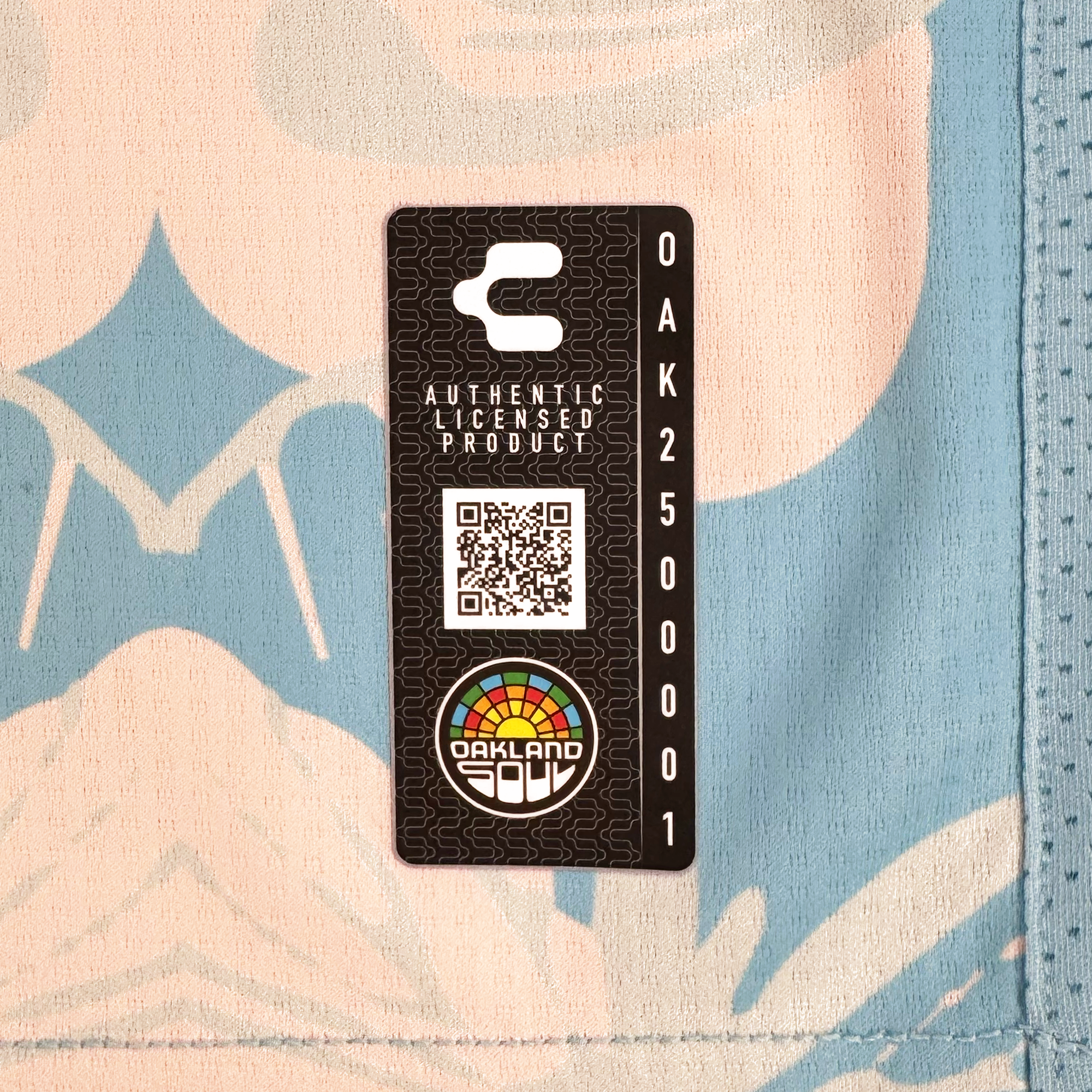 A rectangular black tag with a QR code, “Authentic Licensed Product,” and “OAK 250001” stands on blue and beige patterned fabric. Near the bottom is a rainbow Oakland Soul logo, highlighting Charly's authentic Unisex Butterfly Effect Kit.