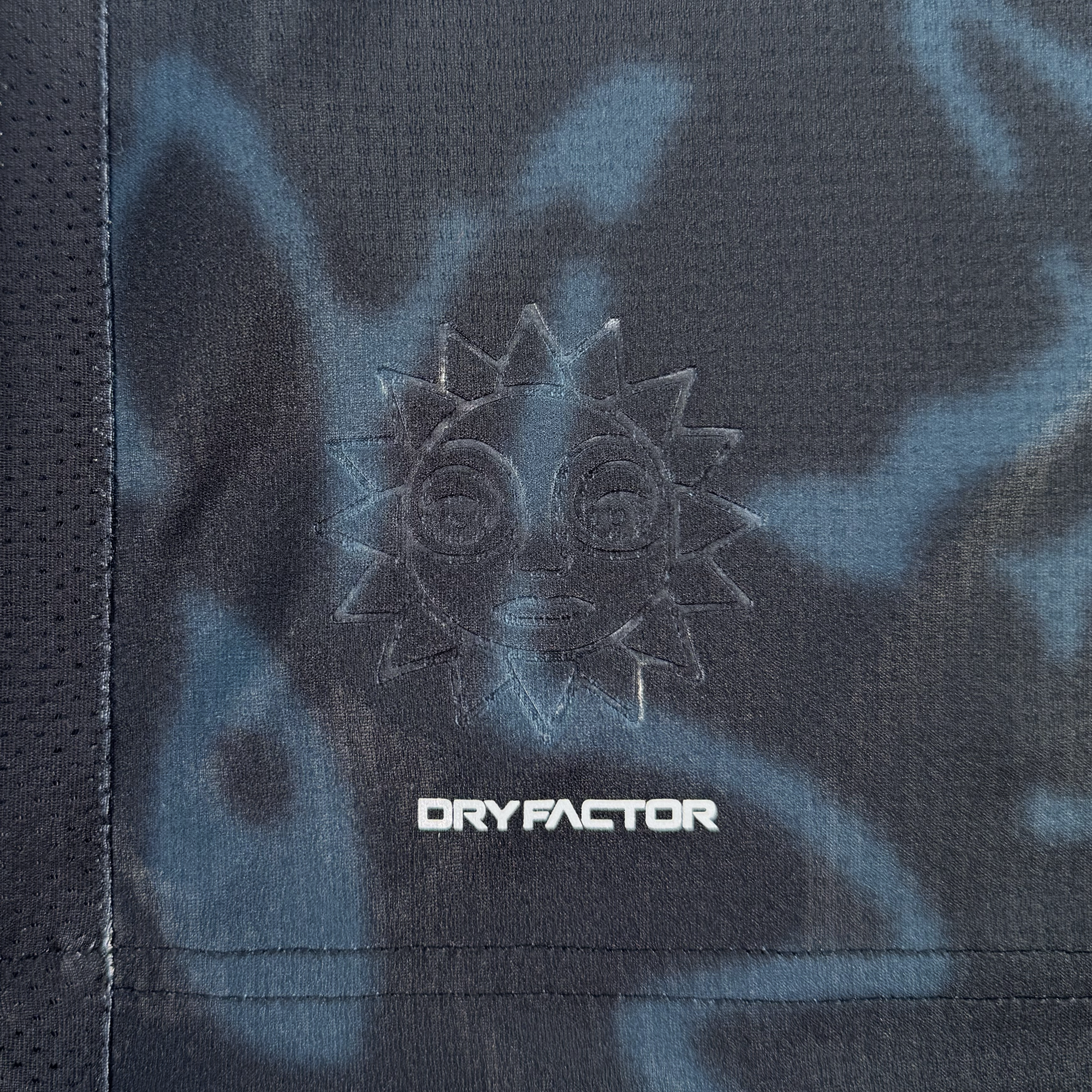 Close-up of a dark Oakland Soul Unisex Soul Bloom Kit fabric with a subtle sun-like embroidered emblem featuring a face. Below the emblem is "DRYFACTOR" in white. The textured technical material has a blue and black floral tie-dye camo pattern.