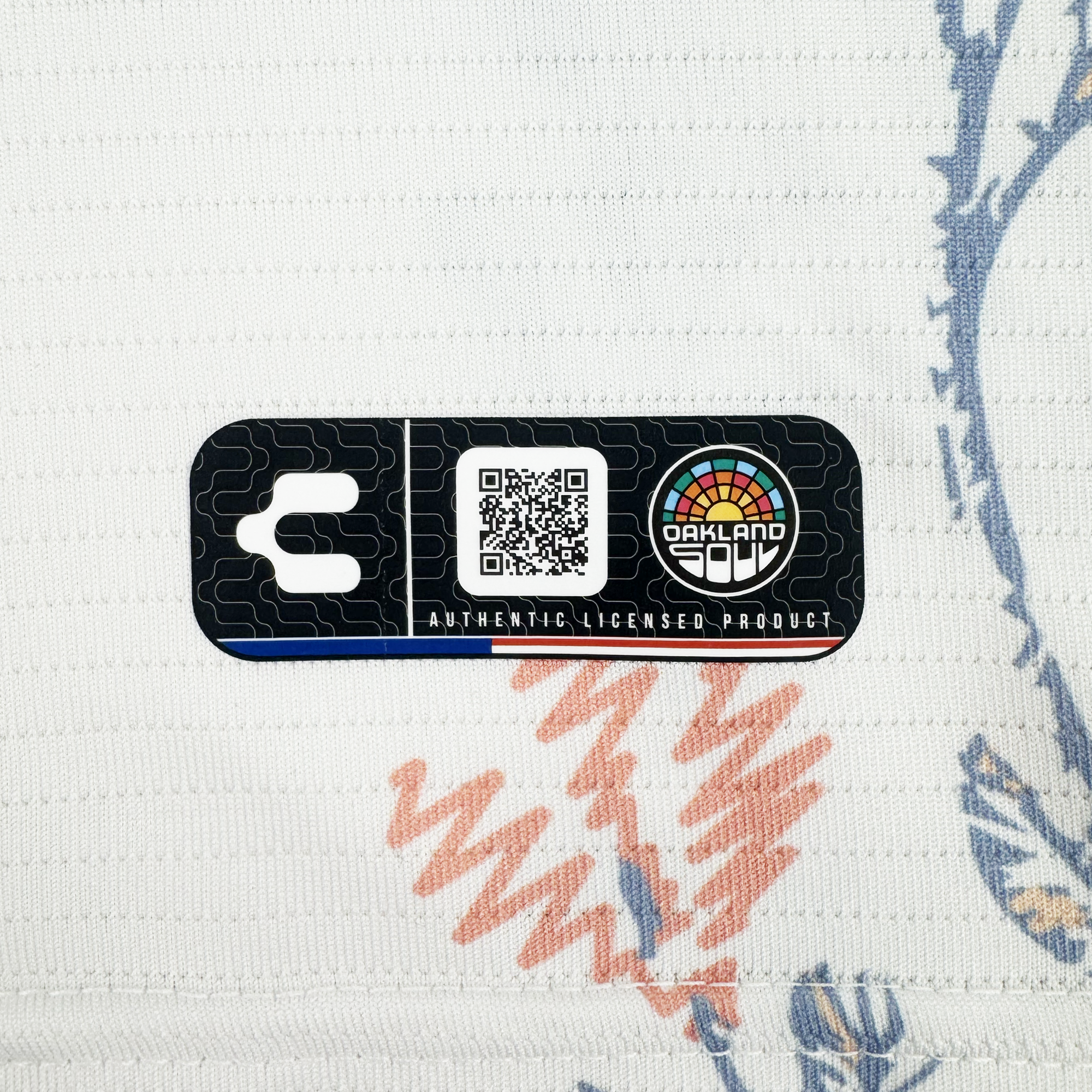 A rectangular black Oakland Soul label with a QR code, “OAKLAND SOUL,” and “AUTHENTIC LICENSED PRODUCT” is sewn onto textured white fabric with blue and red designs on the Custom 2026 OAKLAND SOUL SC FORM JERSEY.