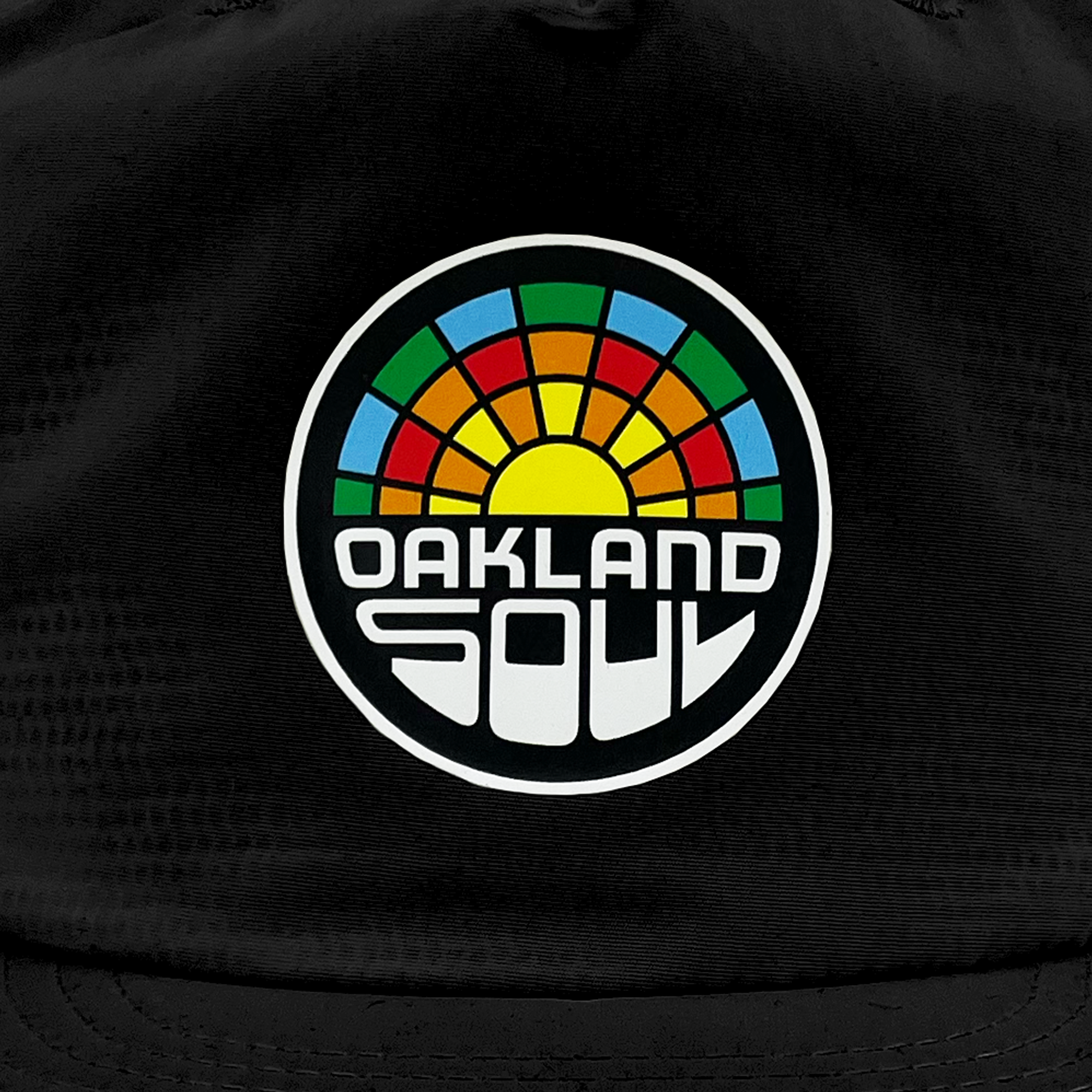 The Oakland Soul Surf Cap features black fabric adorned with a white circular logo, showcasing a multicolored mosaic reminiscent of a sunrise. Beneath the vibrant design, the text "OAKLAND SOUL" is displayed in bold, white, retro-futuristic font. This cap represents Oakland Soul, proudly part of the USL W-League women's soccer team.