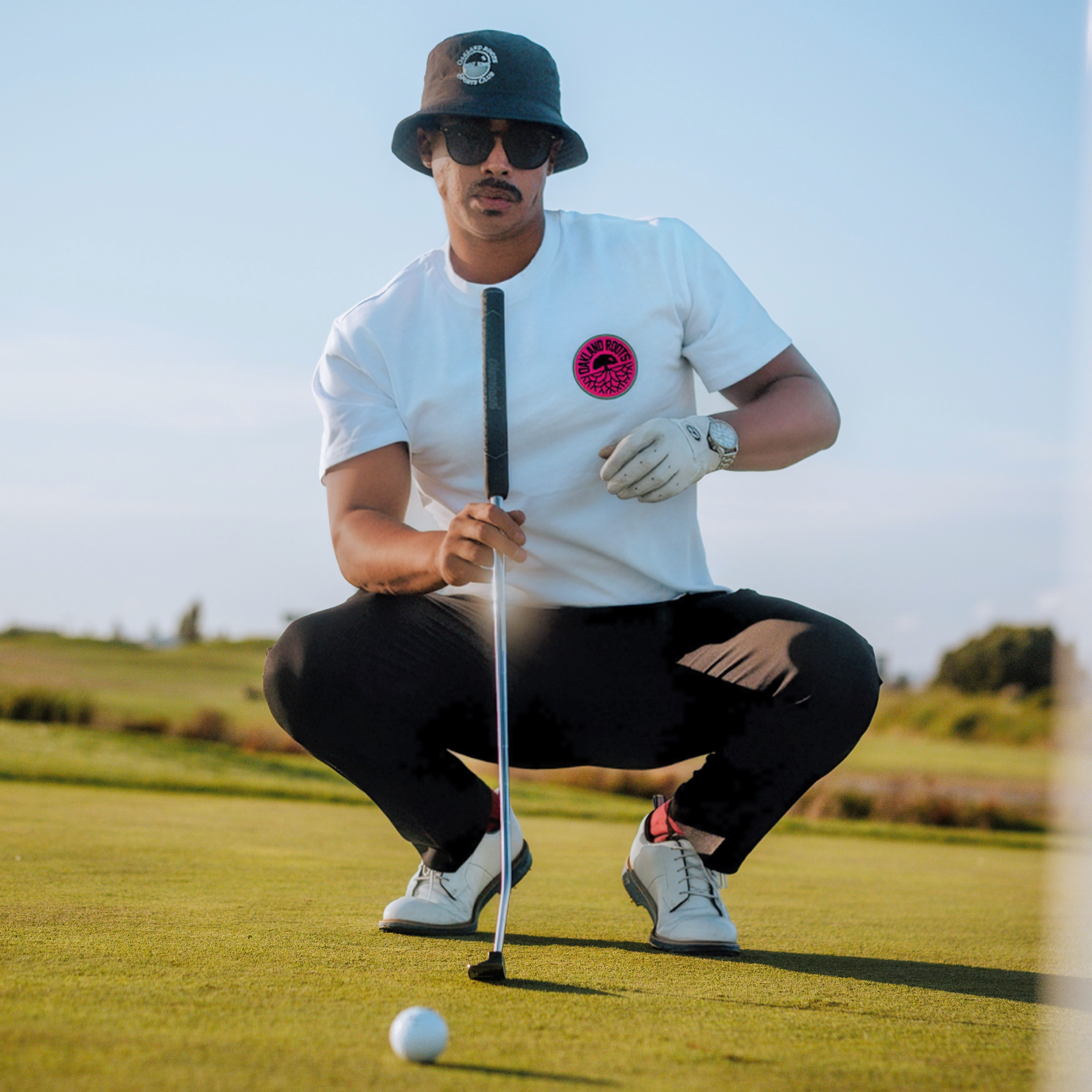 A man squats on a golf course, focusing intently on a golf ball in front of him. He holds a putter and is dressed in an Oakland Roots SC Staycation Heavy Tee featuring a pink and black logo, black pants, white sneakers, and a dark bucket hat. He is also wearing sunglasses and a white golf glove, with a scenic background of grass and trees.