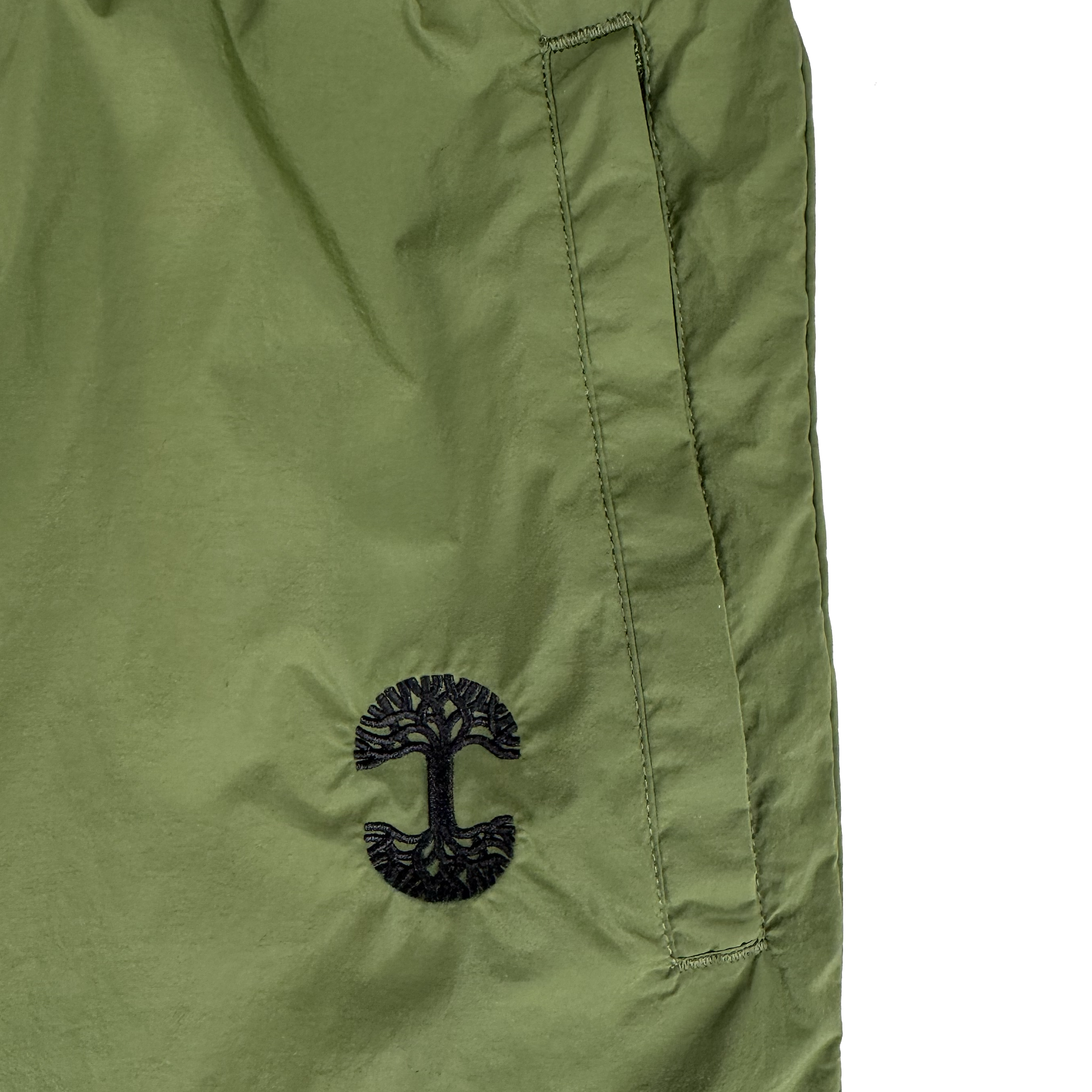 Close-up of Oaklandish's Susurrus Track Bottom in olive green nylon, with a slanted pocket and black tree logo below it. The plain white background highlights its color, texture, and adjustable waistband.