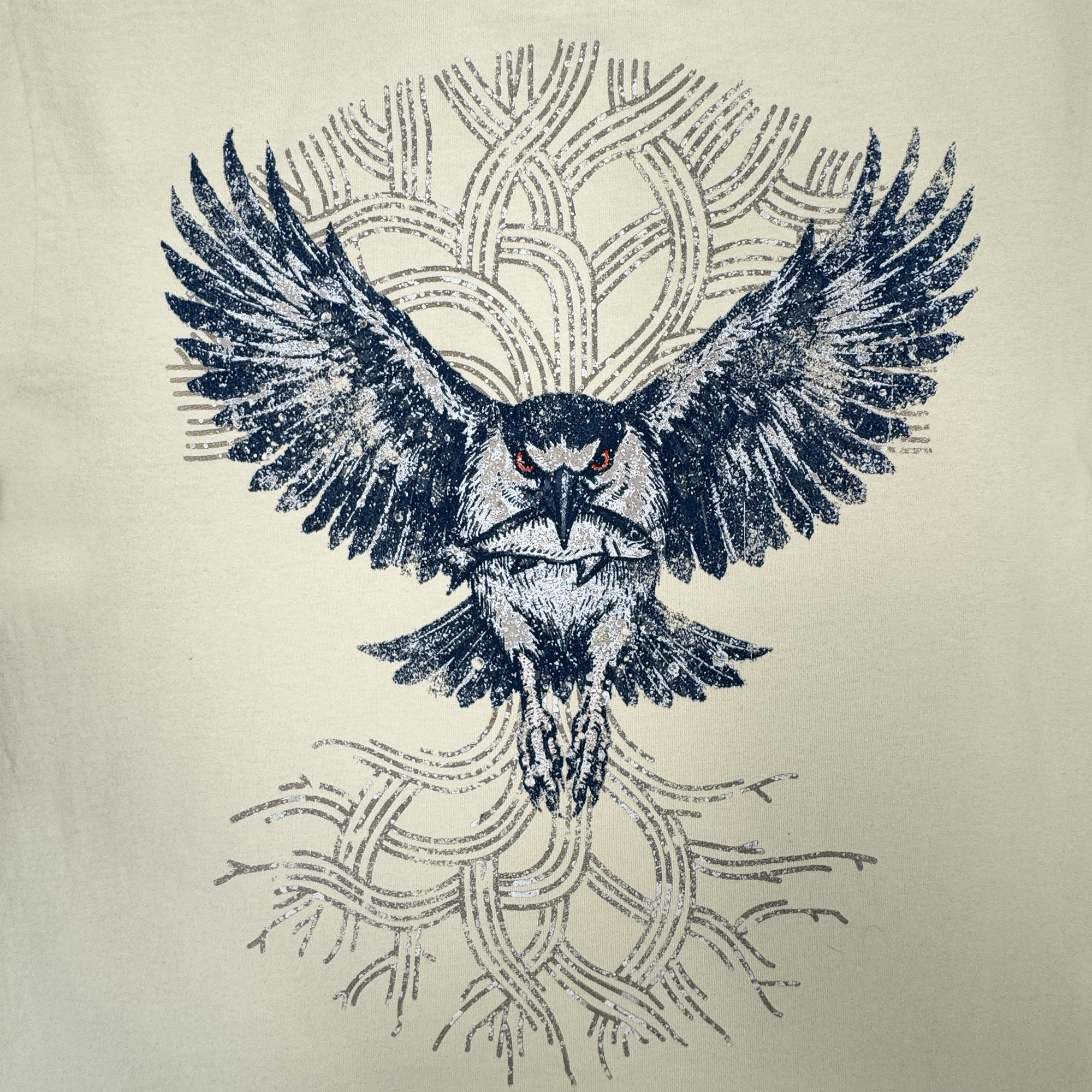 The Oaklandish Swoop Tee features a striking illustration of an owl with outstretched, detailed wings and bold red eyes, swooping across the front. Set against a geometric backdrop of interwoven curved lines in lighter tones, this classic fit men’s t-shirt is crafted from heavyweight light beige cotton for enduring comfort and durability.