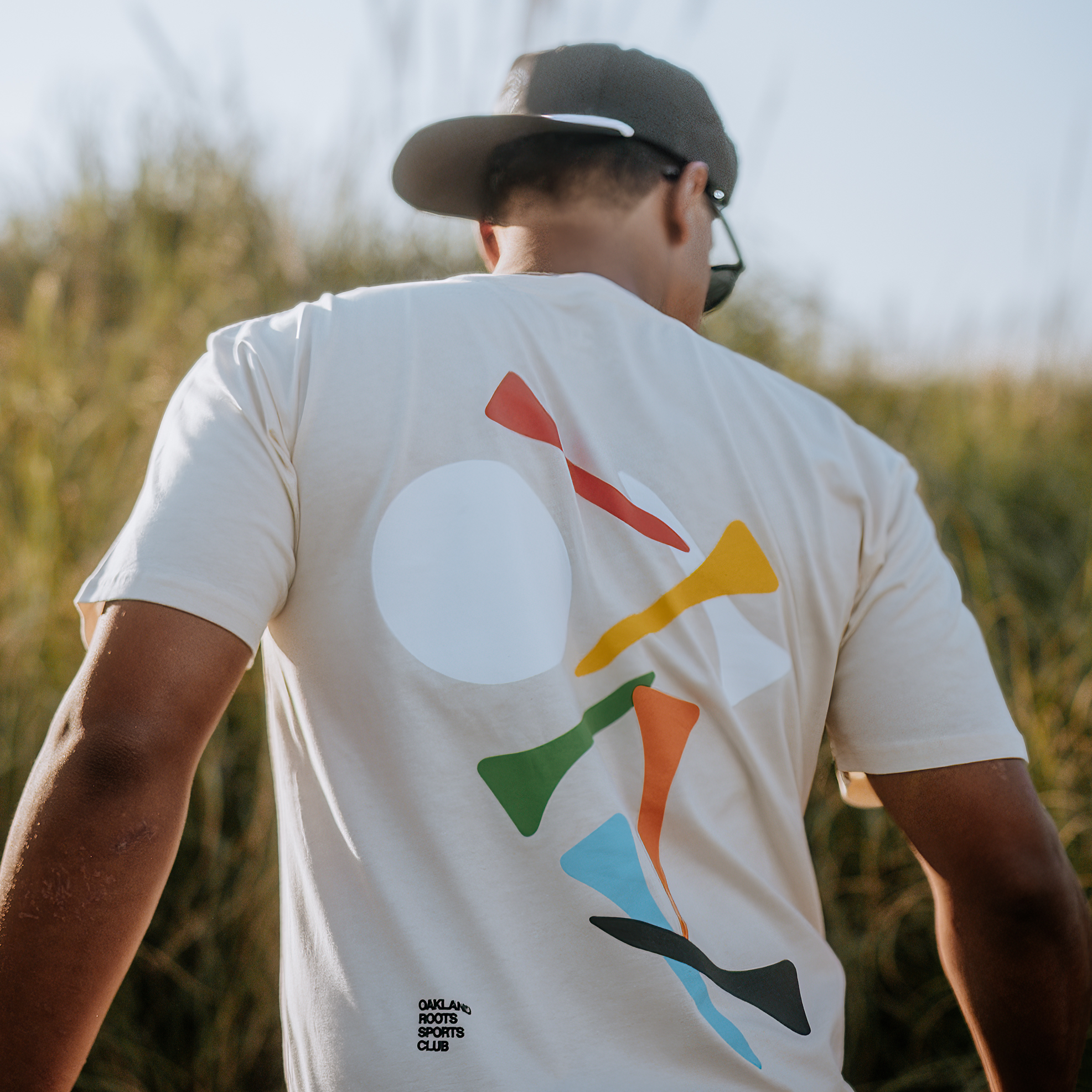 A man faces away, standing outdoors against tall grass. He wears a cap, sunglasses, and a light-colored classic fit Tee Time Tee from Oakland Roots SC. The T-shirt features a circular design with colorful abstract shapes and has the logo "Analog Sports Club" visible on the lower part. The atmosphere is bright and sunny.