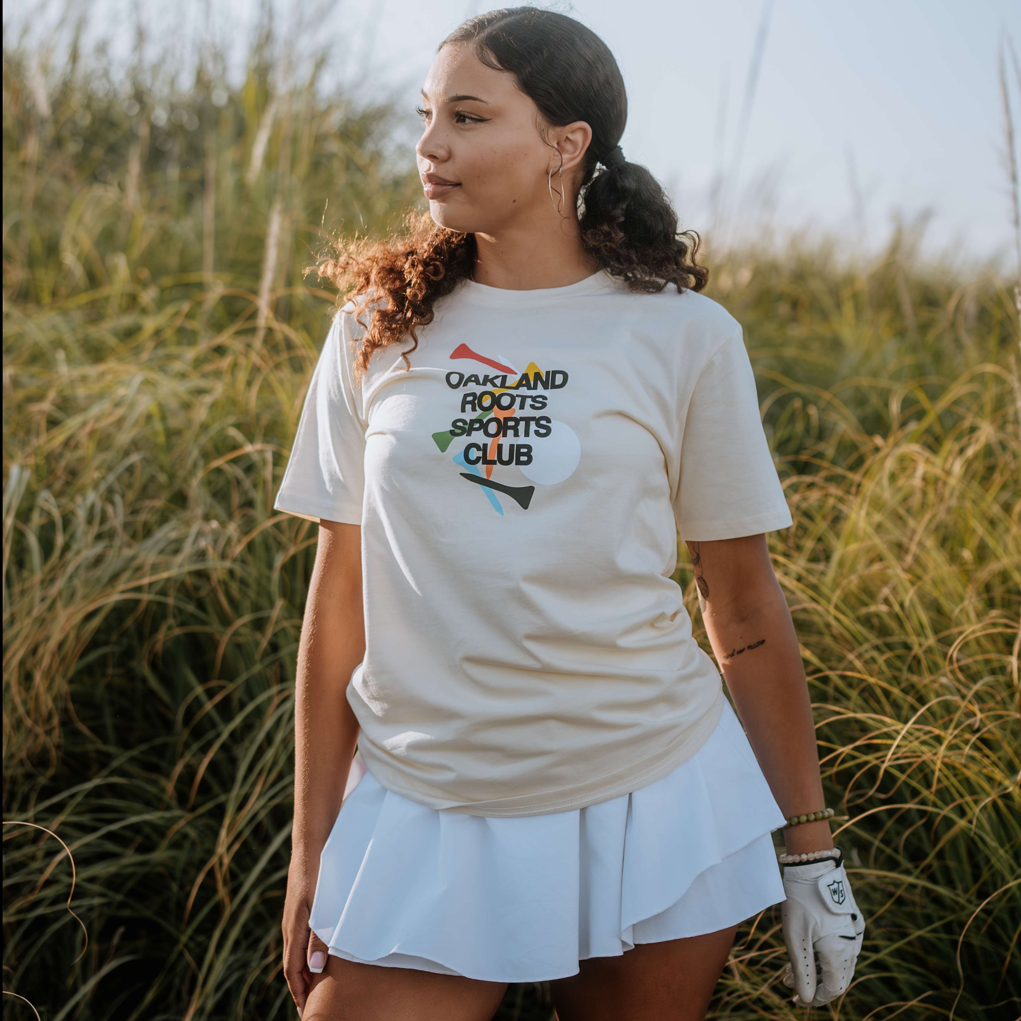 A woman stands outdoors in front of tall grass, wearing the Tee Time Tee from Oakland Roots SC paired with a matching tennis skirt. With her curly hair tied back and hoop earrings, she accessorizes with a white glove on her left hand. The warm, natural lighting underscores her connection to social good through sports club initiatives.