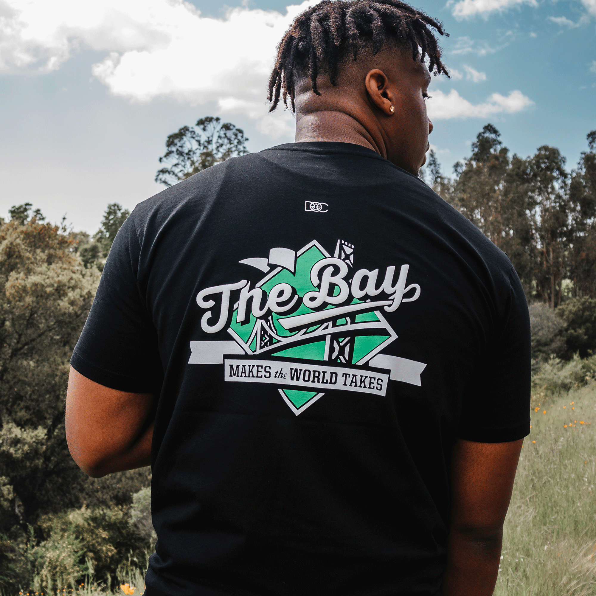 A person stands outdoors, facing away from the camera, wearing "The Bay Tee by DOC" from Oaklandish. The classic fit t-shirt is made of 100% cotton, featuring a design that embodies Bay Area culture with the text "The Bay Makes the World Takes" and images of a bridge, cranes, and buildings in green and white. Trees and a cloudy sky complete the background.