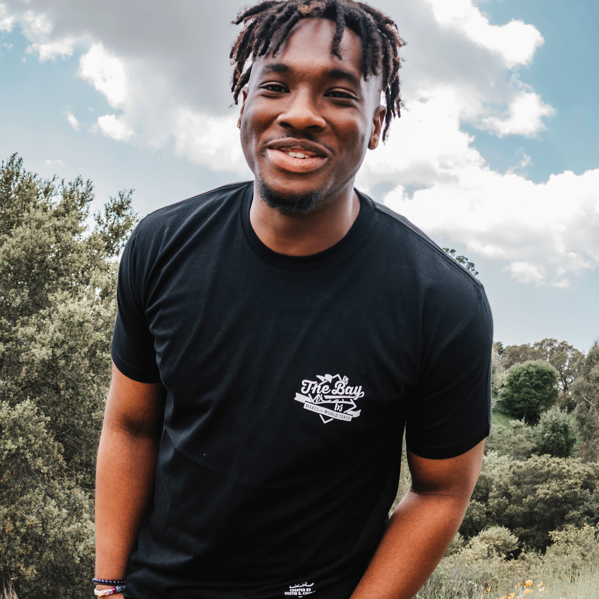 A man with short, twisted hair is smiling broadly while slightly leaning forward. He is wearing The Bay Tee by DOC from Oaklandish, celebrating Bay Area culture. The background features a partly cloudy sky and lush greenery, suggesting an outdoor setting.