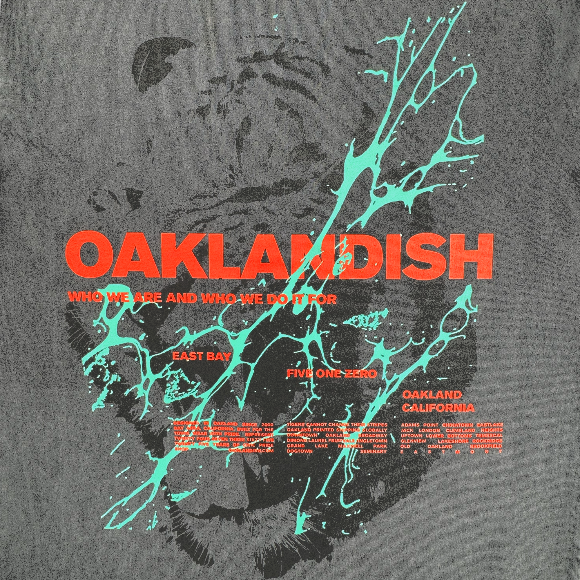 The Tiger Blood Tee by Oaklandish features bold orange-red "OAKLANDISH" text over a grayscale tiger face on a vintage black shirt with turquoise splatters, plus phrases like "Who we are and who we do it for," "East Bay," and "Oakland California.