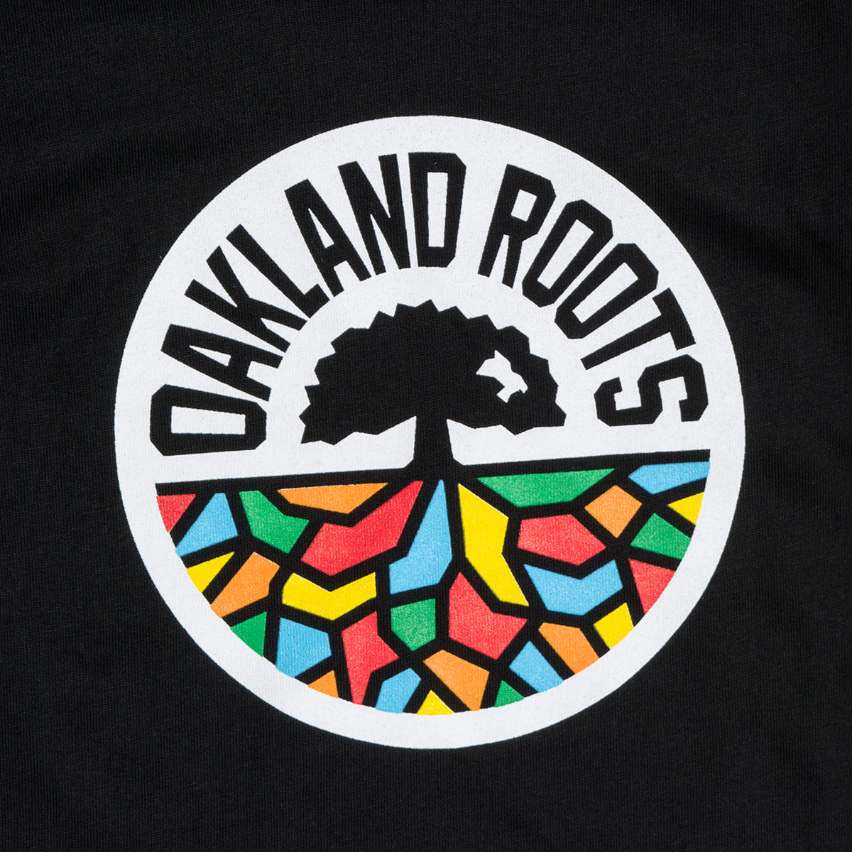 The Toddler Oakland Roots SC Classic Tee showcases the emblem of the Oakland Roots sports club. The design includes a black tree with roots extending downward, creating vibrant geometric shapes in red, yellow, blue, and green. The logo is enclosed within a white circle with "OAKLAND ROOTS" written in bold black letters around the top—ideal for a toddler t-shirt by Oakland Roots SC.