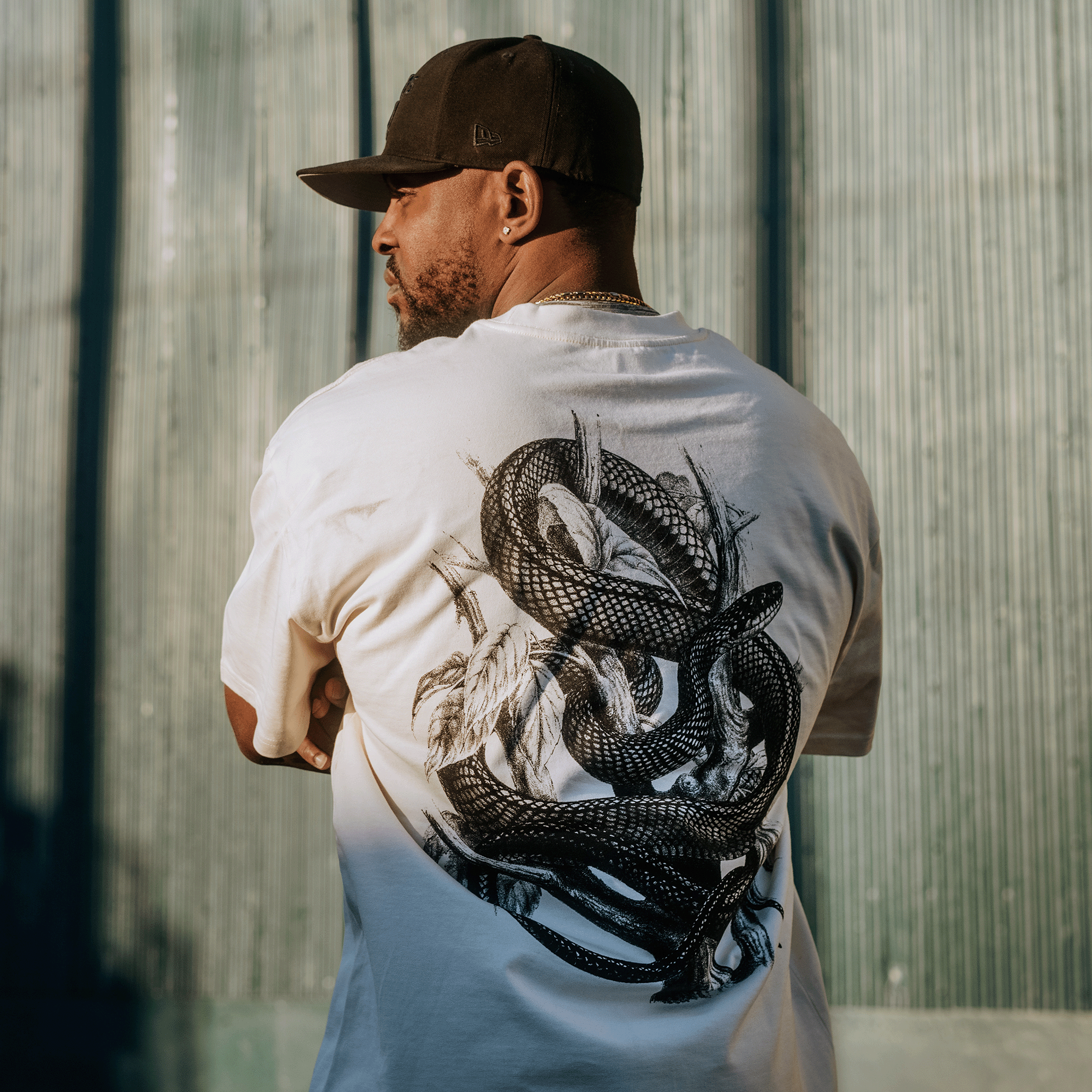 A man stands outdoors with his back to the camera, wearing the Oaklandish Tree Snake Heavy Tee, a boxy fit T-shirt in heavyweight combed cotton. The shirt features a large black snake design entwined with branches and leaves. He accessorizes with a dark baseball cap and chain necklace against a textured wall with vertical green panels.