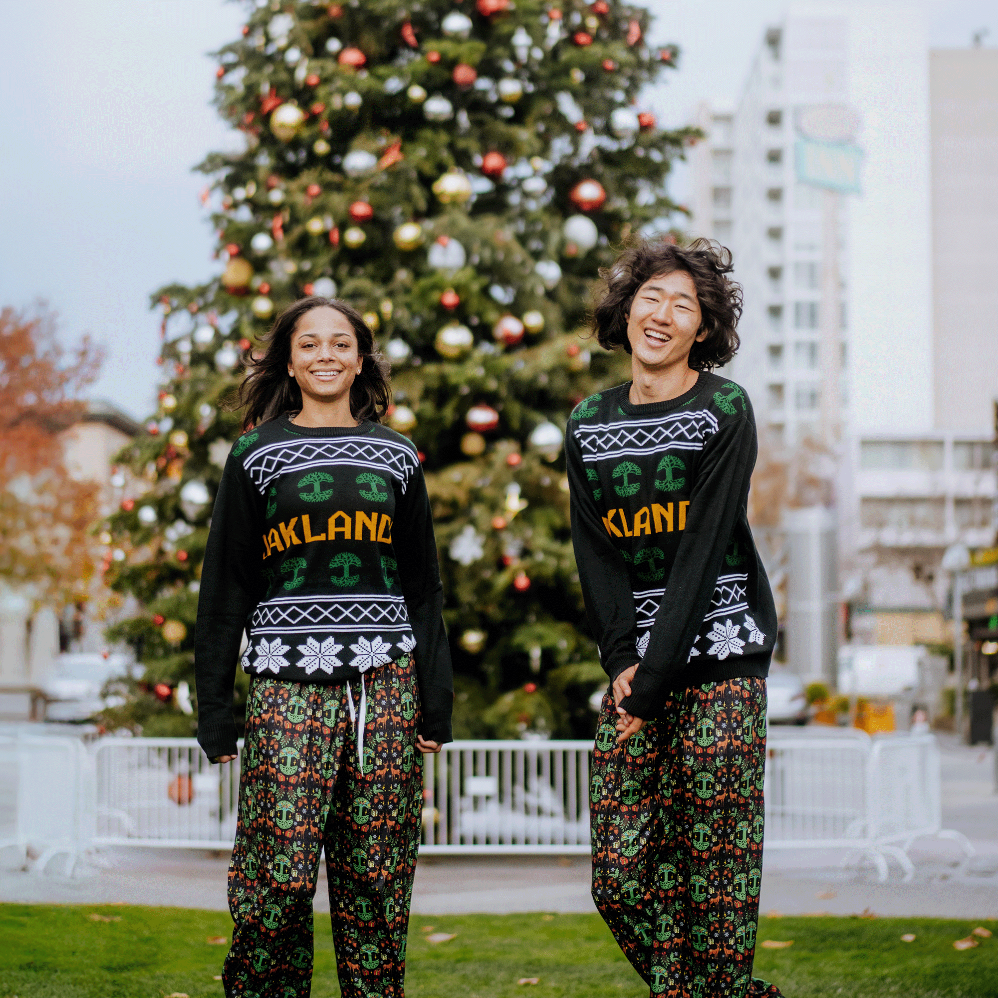 Two people smile in front of a large decorated Christmas tree, wearing matching Oaklandish black sweaters with "Oakland" across the chest and Oaklandish Holiday Pajama Pants. Buildings, a cloudy sky, and white fencing form the background.