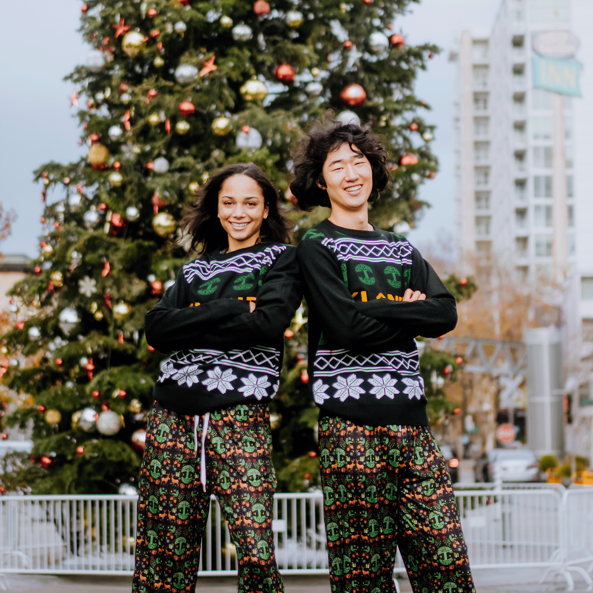 Two people smile with crossed arms in front of a cityscape, featuring a tree and railing. They're wearing matching holiday-themed sweaters and Oaklandish Holiday Pajama Pants by Oaklandish. This creates a cheerful, urban holiday atmosphere.