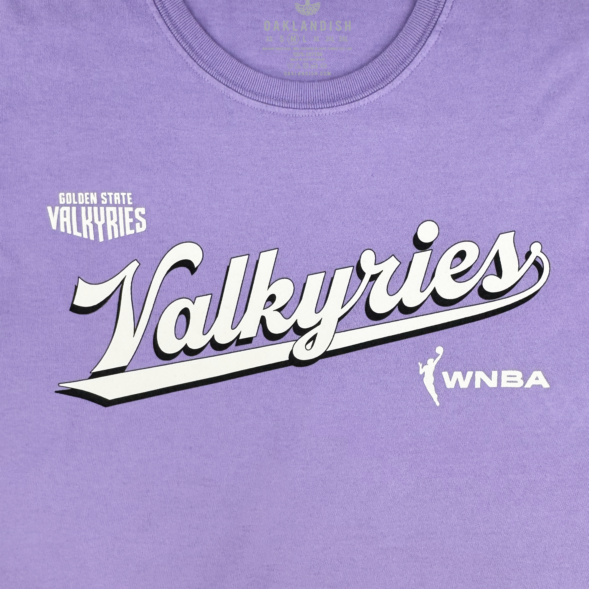 The GSV Golden State Valkyries Retro Script Premium Tee is a lavender organic cotton shirt featuring bold white "Valkyries" script with a black shadow, "Golden State Valkyries" above in small print, and the white WNBA player logo on the right.