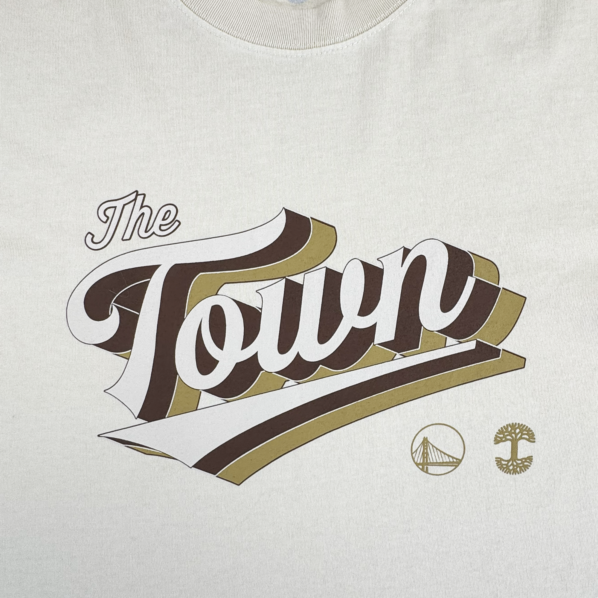 The GSW Vintage Swash Tee is a white shirt featuring "The Town" in bold, brown and gold 3D letters, with Oakland-inspired icons—a bridge and tree—celebrating the Golden State Warriors’ Oakland roots.