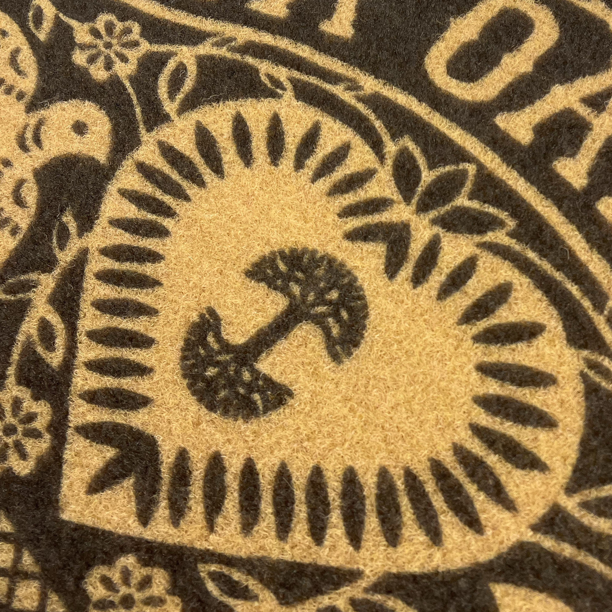 Close-up of a decorative textile pattern on the Viva Oakland Door Mat by Oaklandish, featuring a heart shape with intricate designs. Inside the heart, there is a stylized mushroom graphic. The surrounding area includes floral motifs and partial text, all in contrasting colors of dark and light brown.