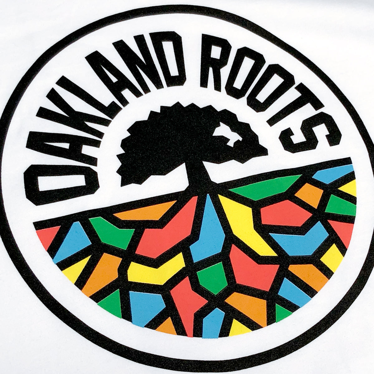 The Women's Oakland Roots SC Classic Tee features a circular logo with "OAKLAND ROOTS" emblazoned in bold black letters, arcing around the top. Beneath the text, a black tree with sprawling roots is illustrated, where the roots form a vibrant mosaic pattern filled with colors such as red, blue, yellow, and green within the lower half of the circle. This emblem captures both the sports club's identity and their dedication to social good.