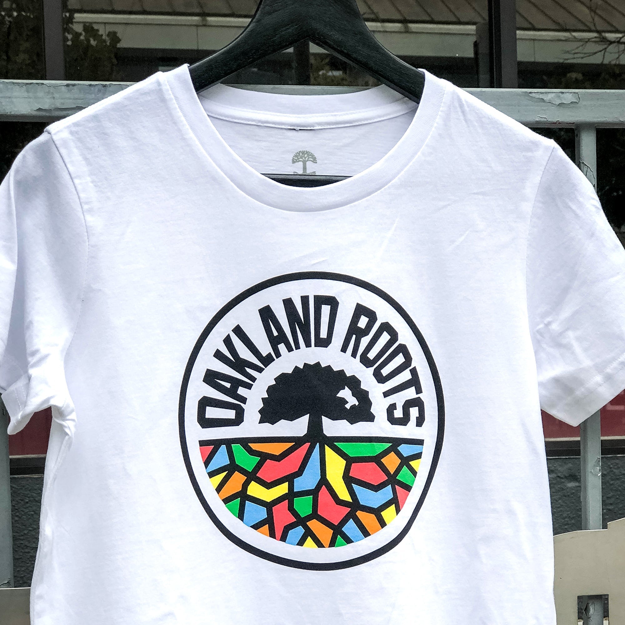 A white "Women's Oakland Roots SC Classic Tee" on a hanger features the logo of the social good-focused sports club, "Oakland Roots SC." The logo includes a black silhouette of a tree and roots, surrounded by the words "Oakland Roots." Below the tree, the roots are depicted in colorful geometric patterns. In the background, there is a metal railing and a window.