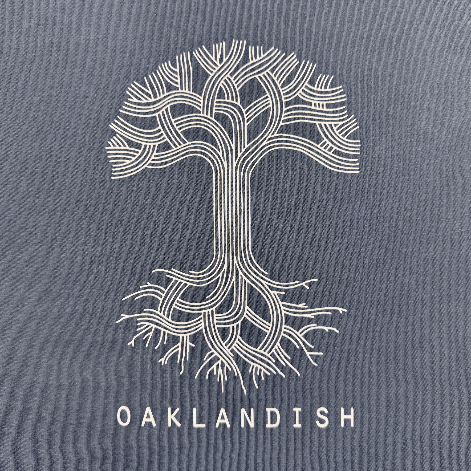 The Women's Oaklandish Classic Logo Tee by Oaklandish features a stylized tree with intricate branches and roots on a classic fit t-shirt. "OAKLANDISH" appears in bold white letters, crafted from 100% cotton with clean lines against faded blue fabric.