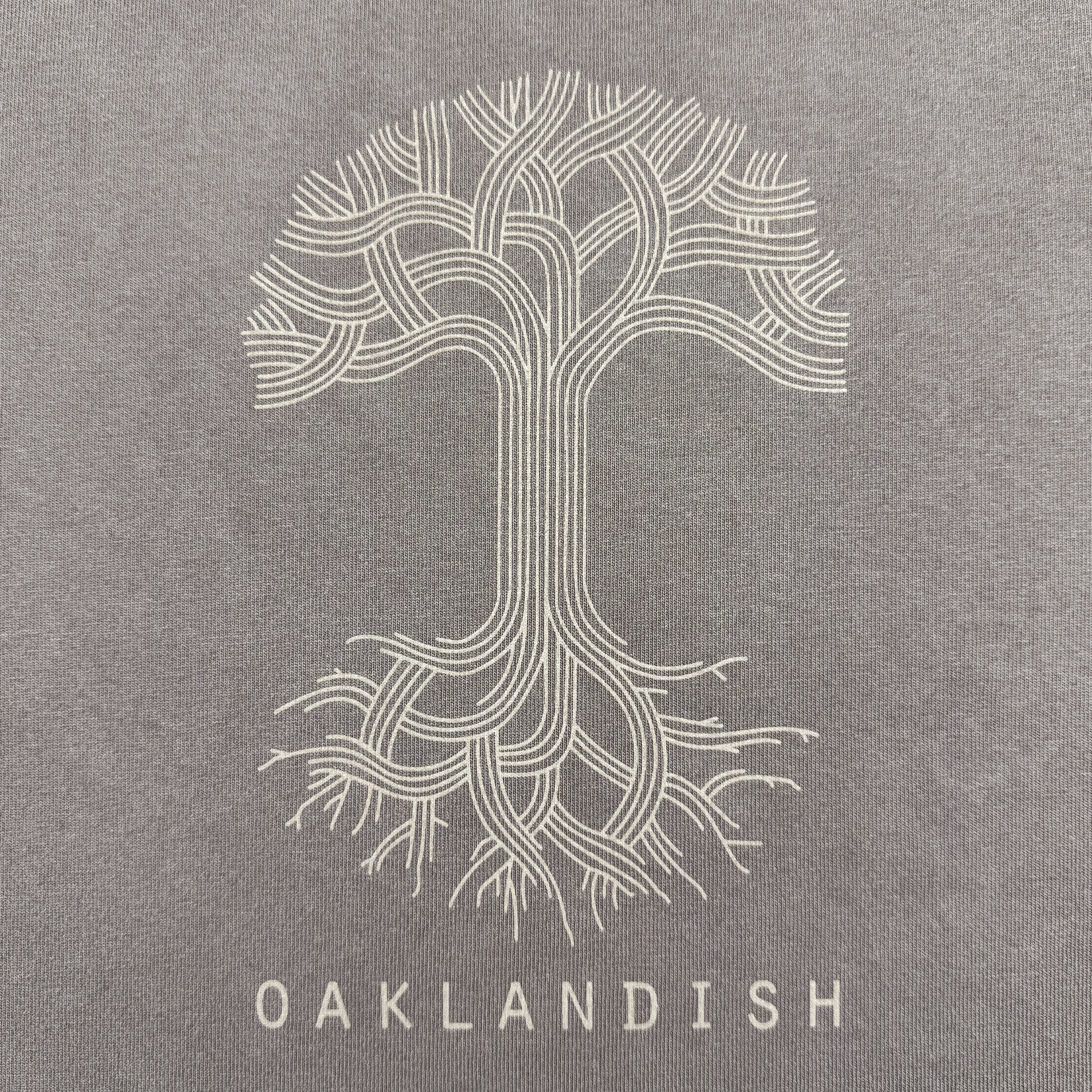 The Women's Oaklandish Classic Logo Heavy Tee from the Oaklandish brand features a stylized tree with interwoven branches and roots on a faded grey oversized fit shirt, with "OAKLANDISH" printed in white below. The symmetrical, line-based design offers a modern artistic look on 100% heavy-weight cotton.