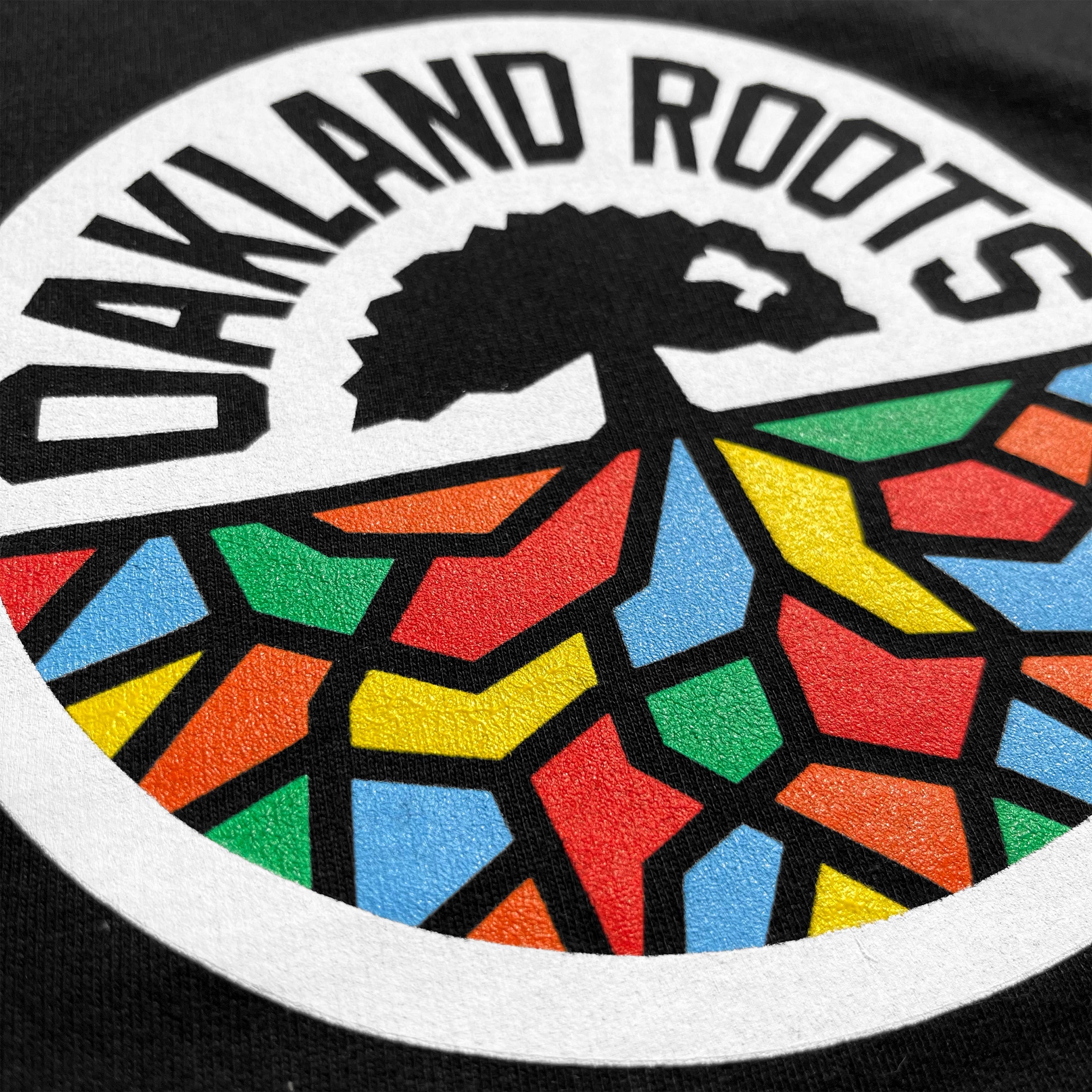 A circular logo with "OAKLAND ROOTS" in bold, white uppercase letters along the top. The bottom half features a tree with sprawling roots, filled with a colorful mosaic pattern in red, blue, green, and yellow on a black background. Perfect for showcasing on the Youth Oakland Roots SC Hoodie by Oakland Roots SC.