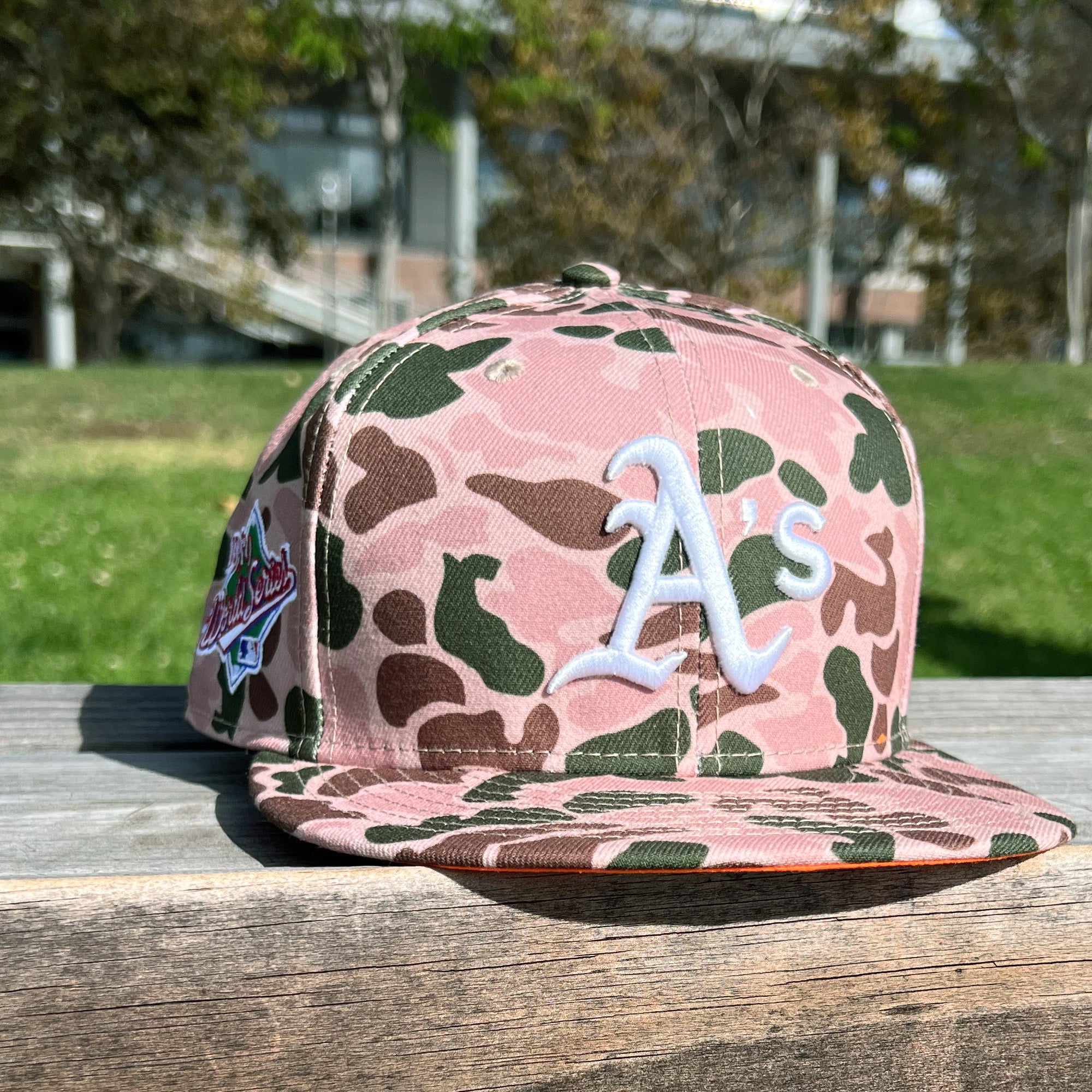 The New Era Oakland A's 59FIFTY Fitted Cap from Retail Vendors, featuring a pink and green Duck Camo pattern, rests on a wooden surface outdoors. The front displays a white "A's" logo, and there's a patch on the right side of the fitted cap. In the background, you can see green grass, trees, and a blurred building structure.