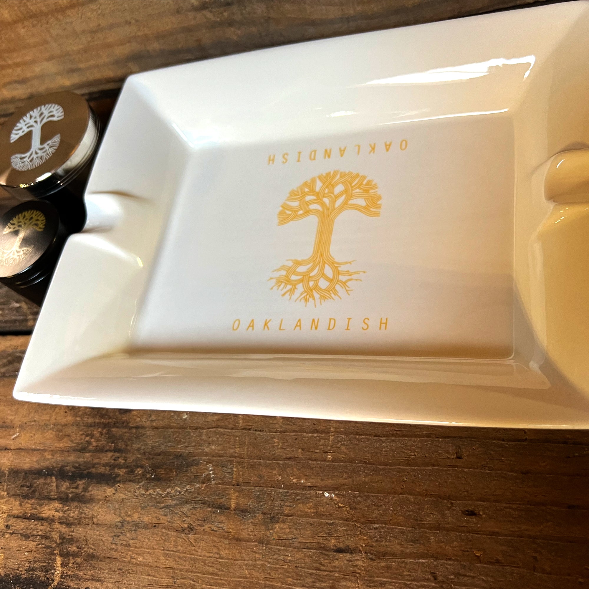 The Oaklandish Ceramic Ashtray, a white rectangular dish with raised edges, displays a central emblem of a yellow tree with the word "OAKLANDISH" above and below it in yellow text. This ceramic imprinted ashtray rests on a wooden surface, with two black and silver round containers partially visible in the background.