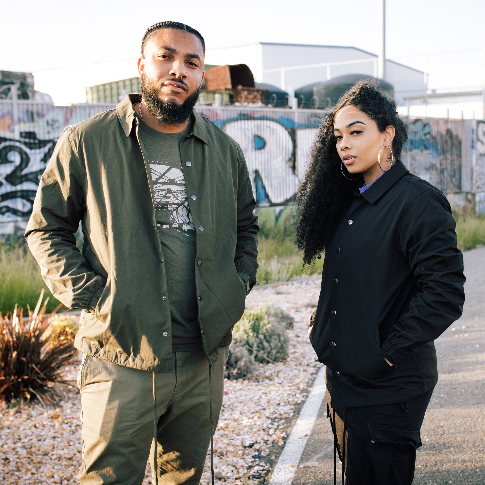 Two individuals stand side by side outdoors in front of a colorful graffiti-covered wall. The person on the left wears a green, water-resistant jacket and pants, while the person on the right sports an Oaklandish Premium Coaches Jacket and black pants. Both have serious expressions and casual postures against an industrial backdrop.