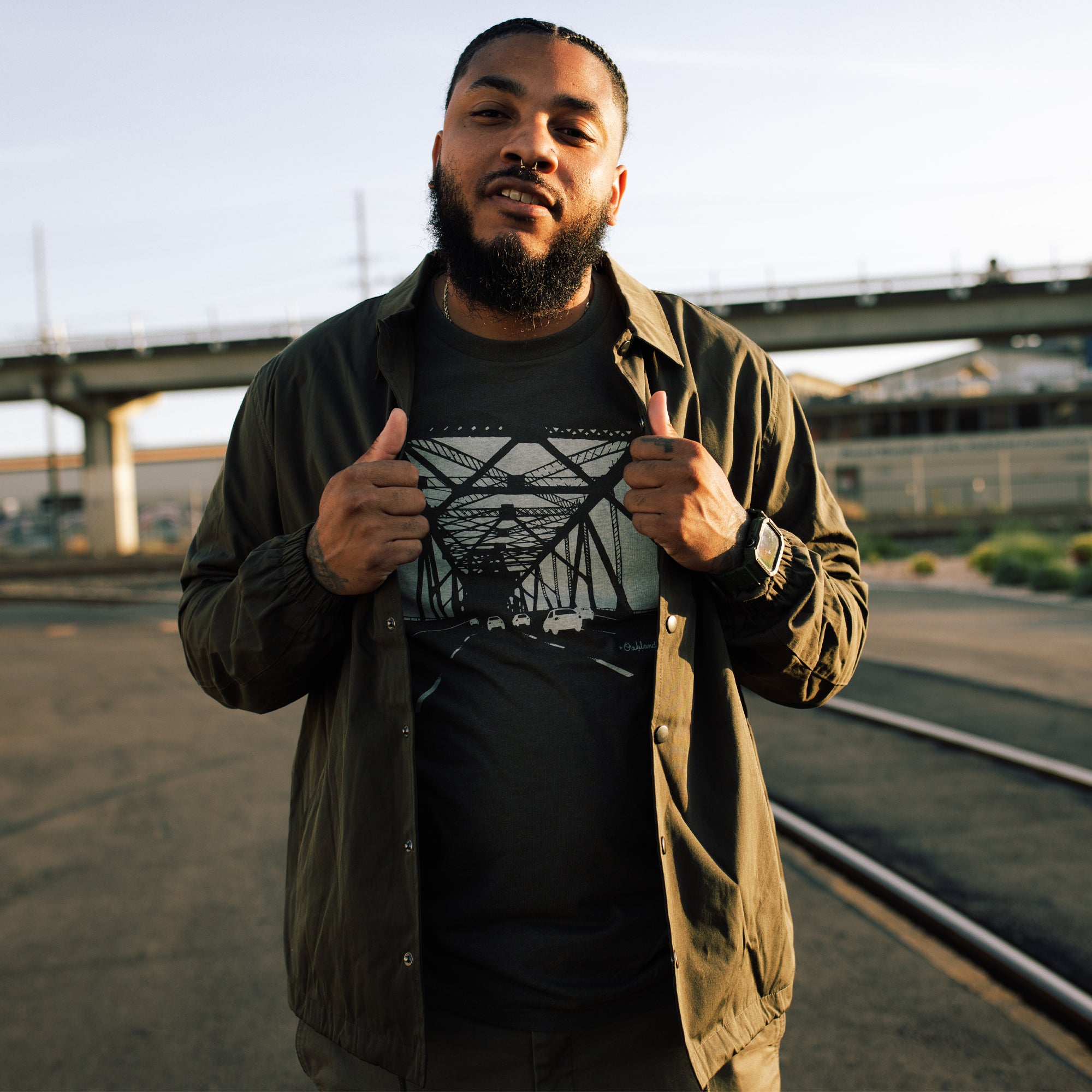 A man with a beard stands confidently outdoors, grasping the open sides of his olive-green, water-resistant Premium Coaches Jacket by Oaklandish to reveal a t-shirt underneath. The t-shirt features an intricate geometric design. Behind him, there is a bridge and railway tracks. The setting sun casts a warm glow on the scene.