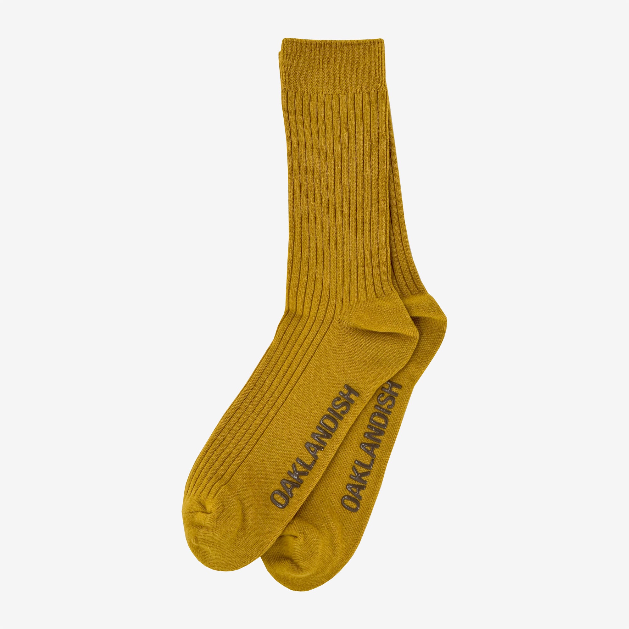 A pair of mustard yellow Grip Crew Socks by Oaklandish, featuring "OAKLANDISH" in dark gray capital letters along the sides. These yellow accessories have a ribbed texture from top to ankle, with reinforced toe and heel areas slightly darker than the rest. They are made from a cotton polyester spandex blend.