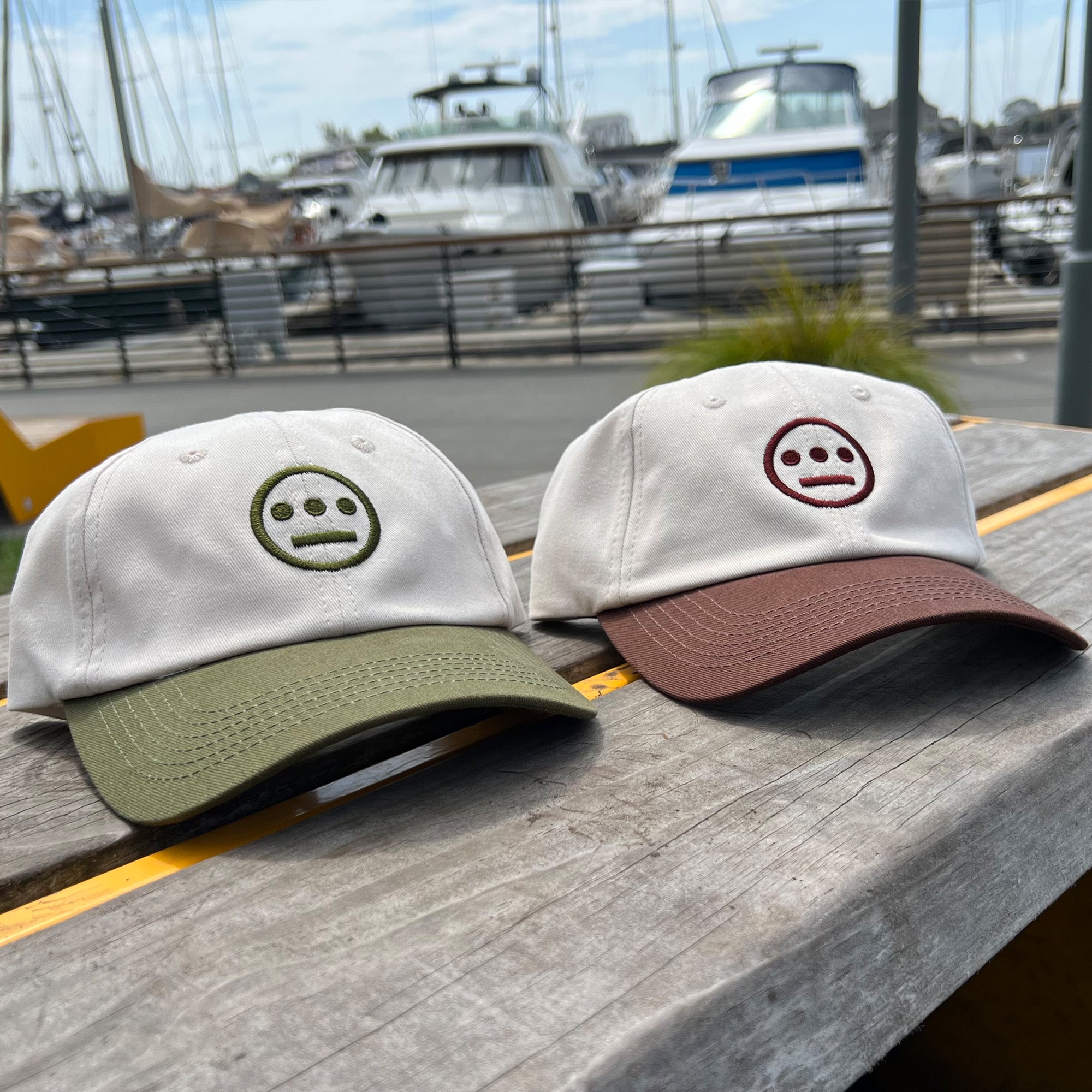 Two Hieroglyphics Hiero Logo Dad Hats rest on a wooden surface by a marina. The hat on the left features a white crown, green brim, and an embroidered unhappy face in green. The hat on the right boasts a white crown, brown brim, and an embroidered unhappy face in burgundy. Boats and a dock are visible in the background.