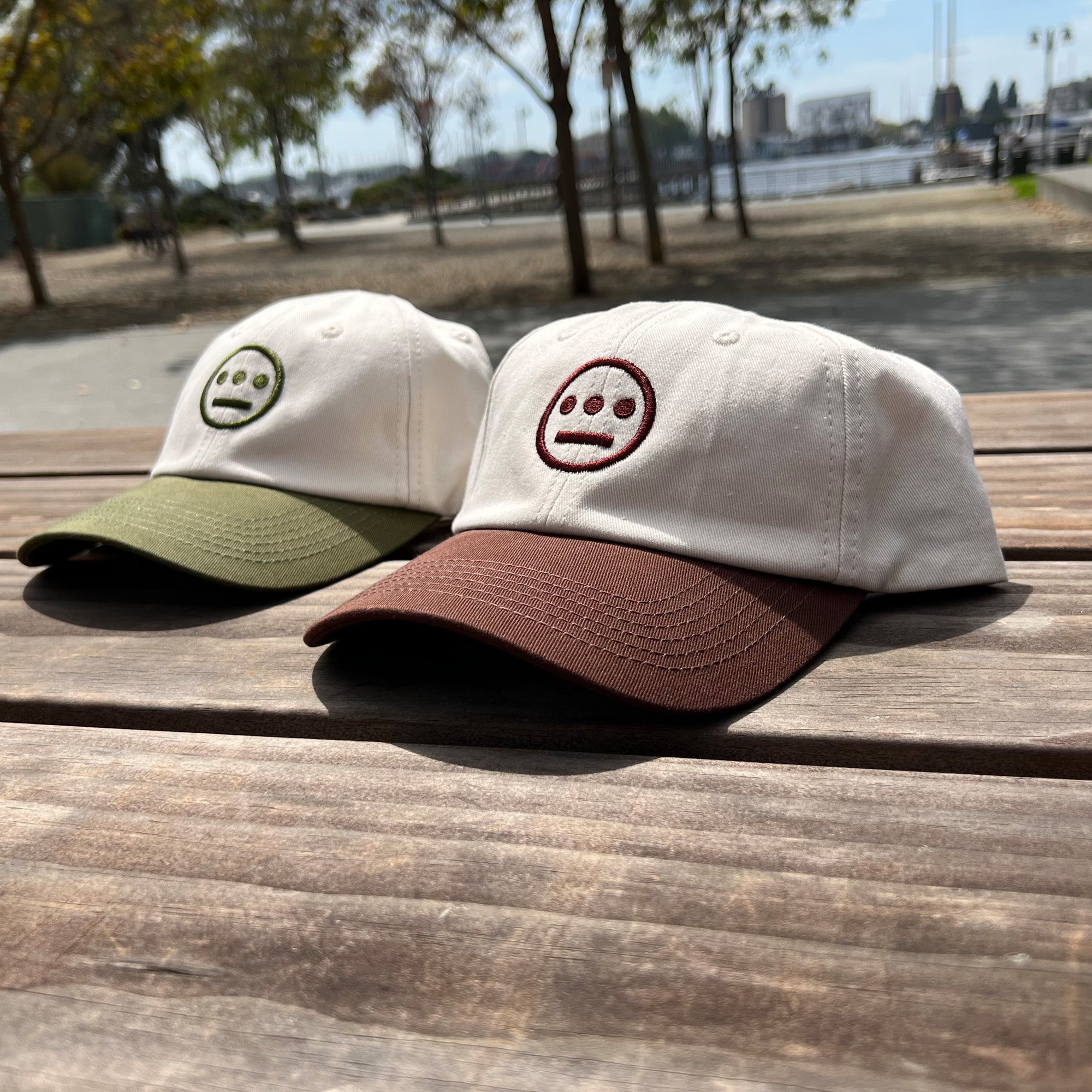 Two Hiero Logo Dad Hats from Hieroglyphics are placed on a wooden table outdoors. Both caps are white with curved brims; one has a brown brim and the other green. Each hat features a stitched design of a simple face with three eyes and a wide mouth, resembling an emoji-like hieroglyphic. Trees and a waterfront are visible in the background.