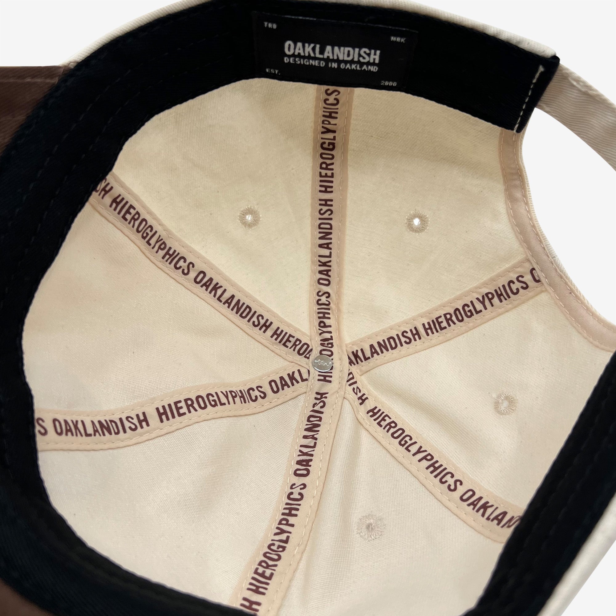 Top view of the interior of the Hiero Logo Dad Hat by Hieroglyphics, showcasing its beige and black design. The inner seams feature repeating "OAKLANDISH" and "HIEROGLYPHICS" in black capital letters. A central label reads "OAKLANDISH DESIGNED IN OAKLAND." The hat includes ventilation holes and a black band.